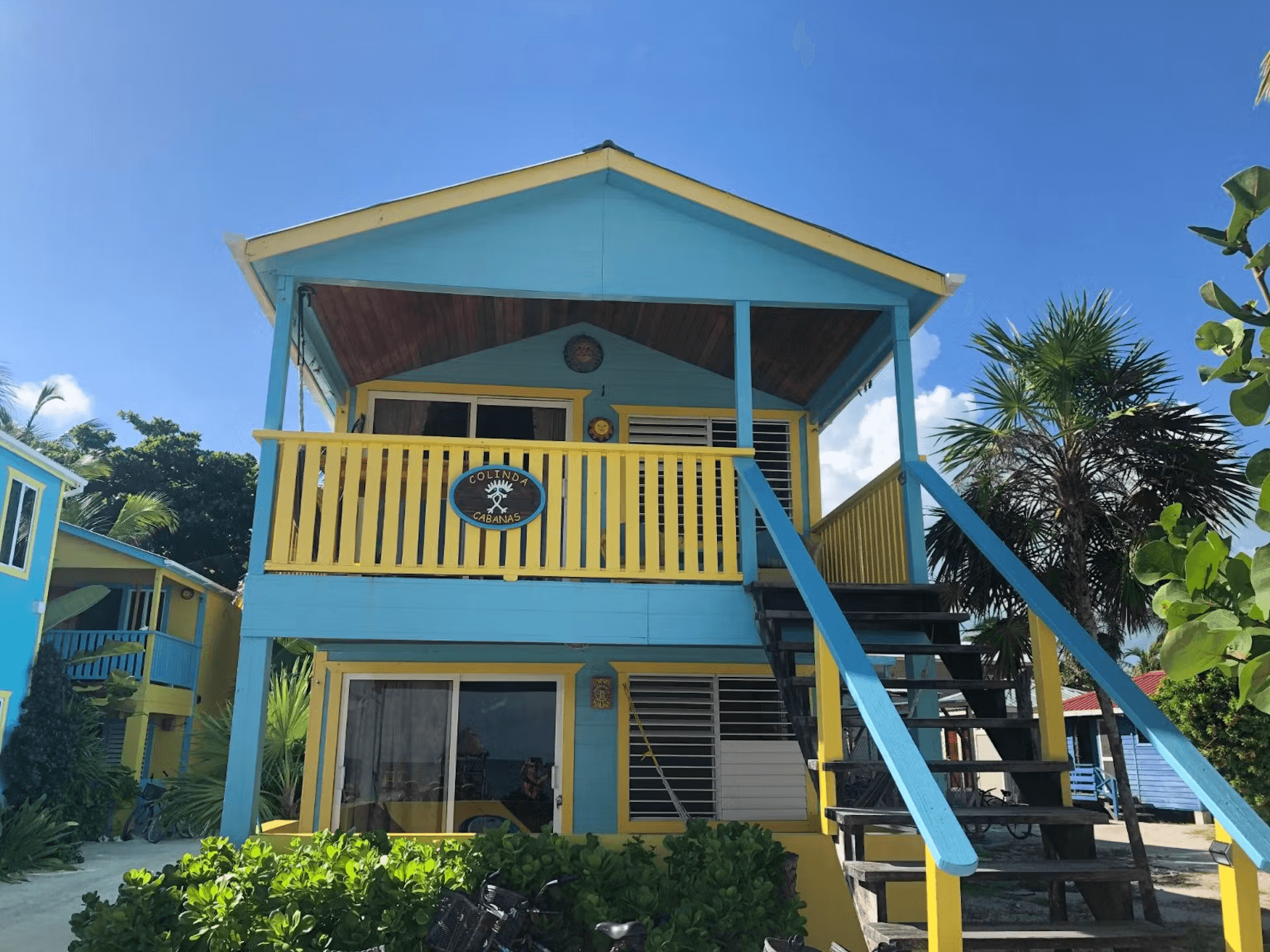 Exterior view of a colorful blue and yellow cabana with stairs leading to second level, bright blue sky and tropical foliage