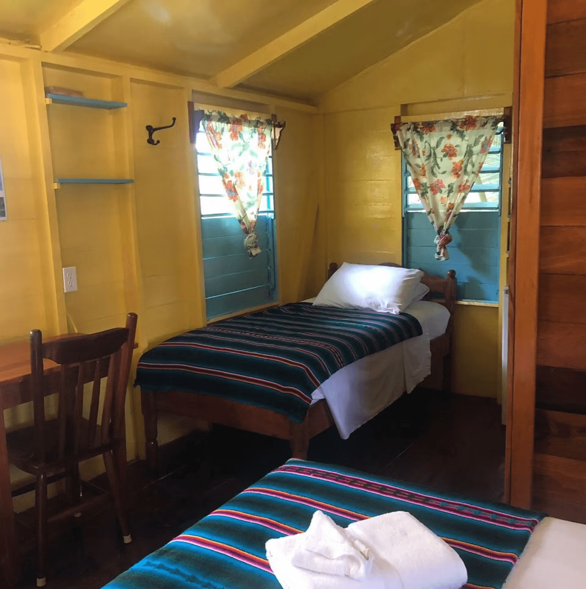 Bedroom with yellow painted walls, showing foot of double bed with single bed in opposite corner, wood table and matching chair, two sunny windows with blinds and curtains
