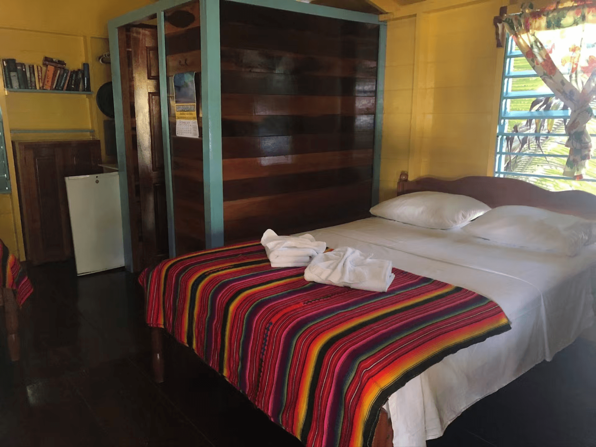 Bedroom with bright yellow walls, hardwood accents, and double bed with colorful striped blanket and white sheets
