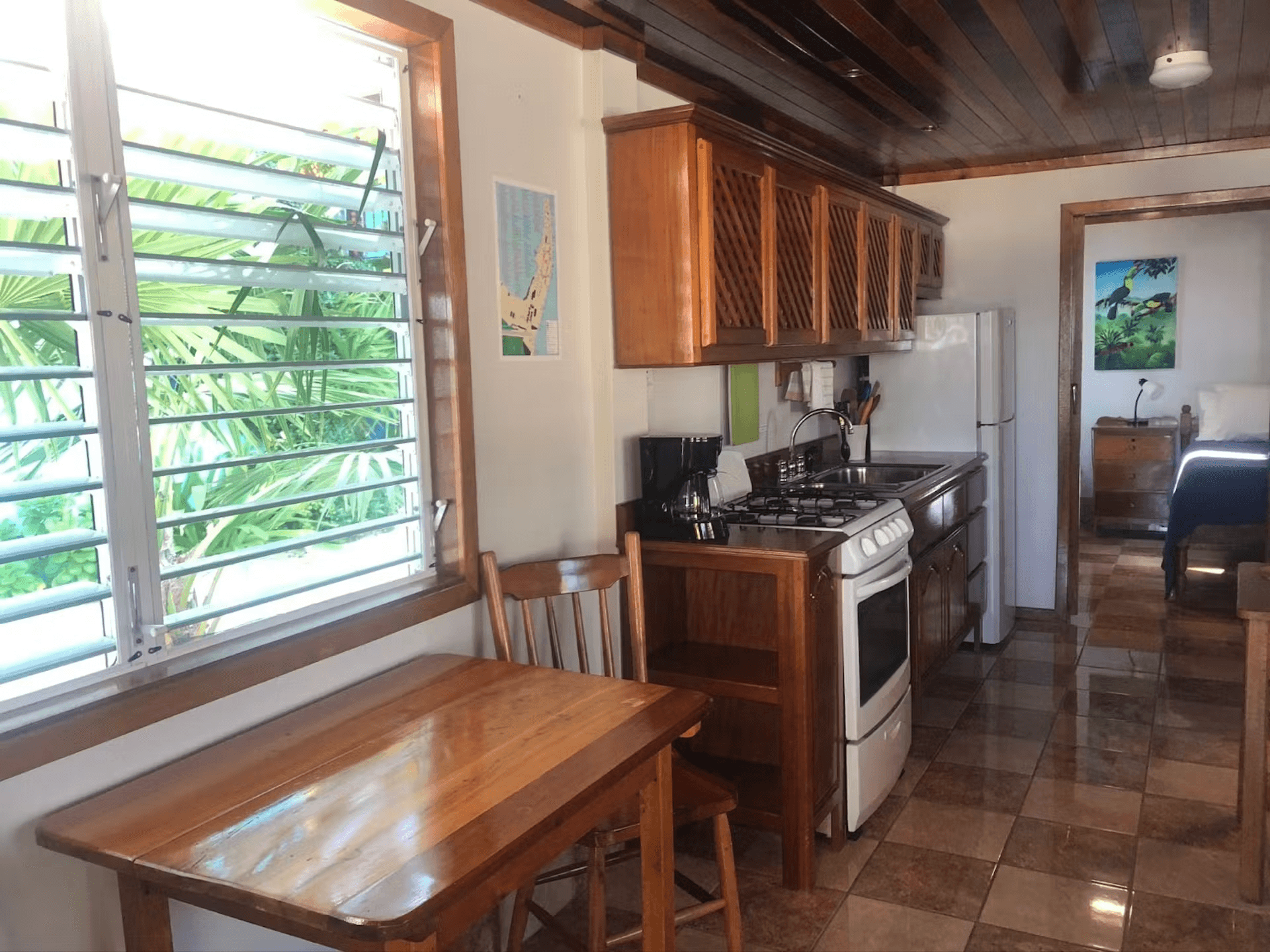 Kitchen table and chairs under window, kitchen cabinets, stove and refrigerator, and doorway leading to bedroom