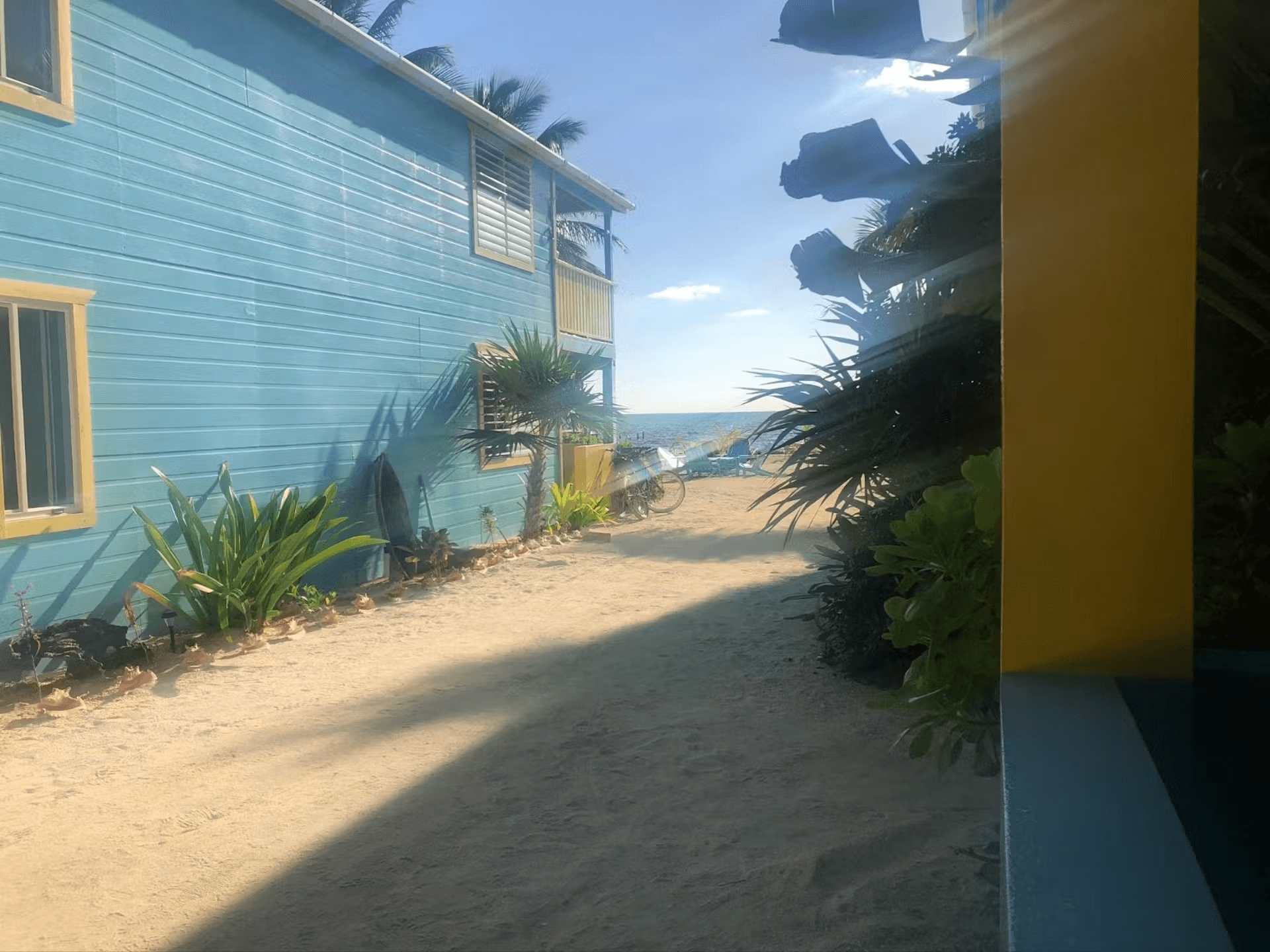 The sun streams between two brightly painted cabanas separated by sand and tropical foliage