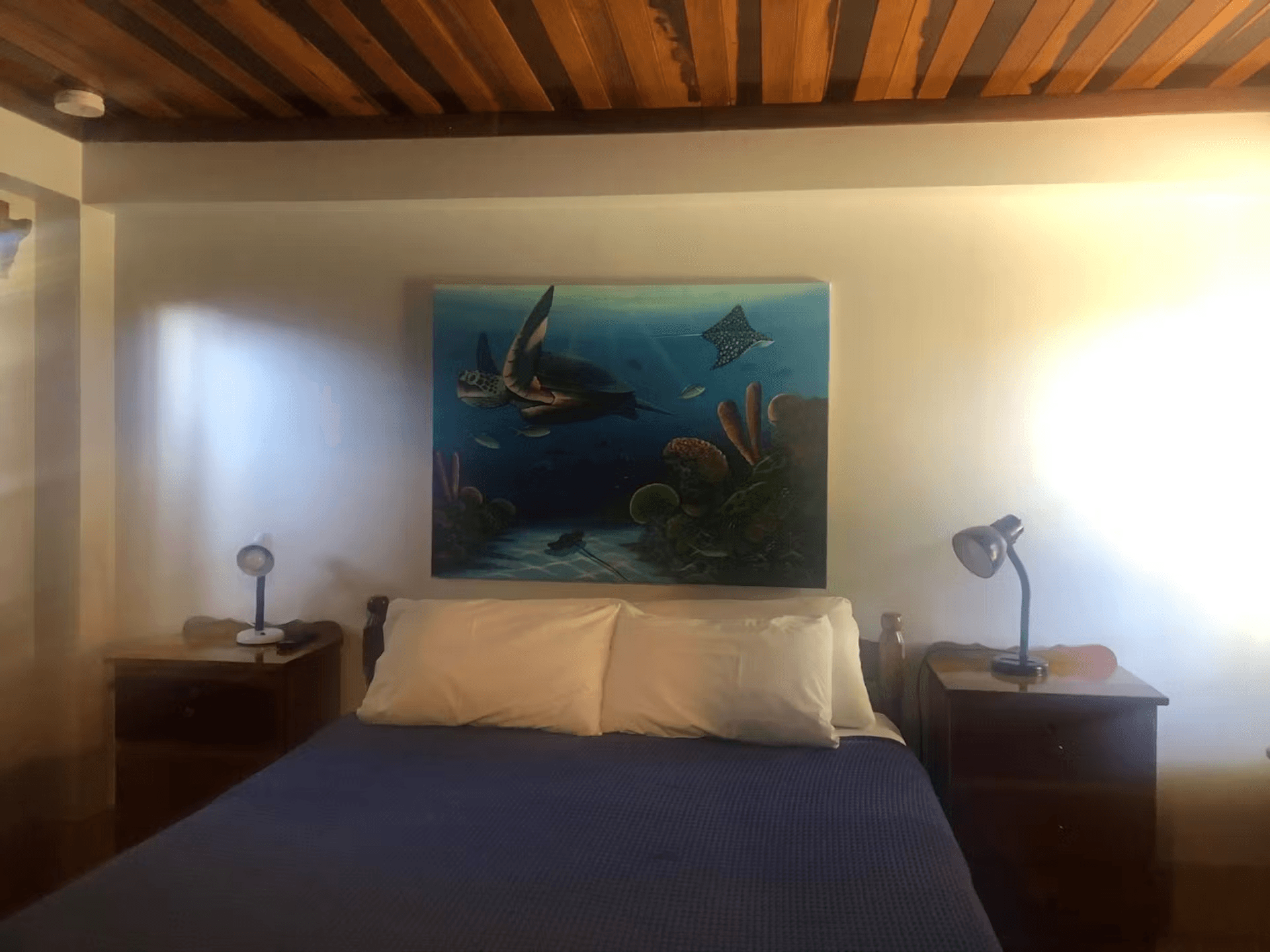 Bedroom with queen bed made with blue bedspread and white sheets & pillowcases, two nightstands with table lamps, and ocean-themed artwork hangs on the wall above the bed