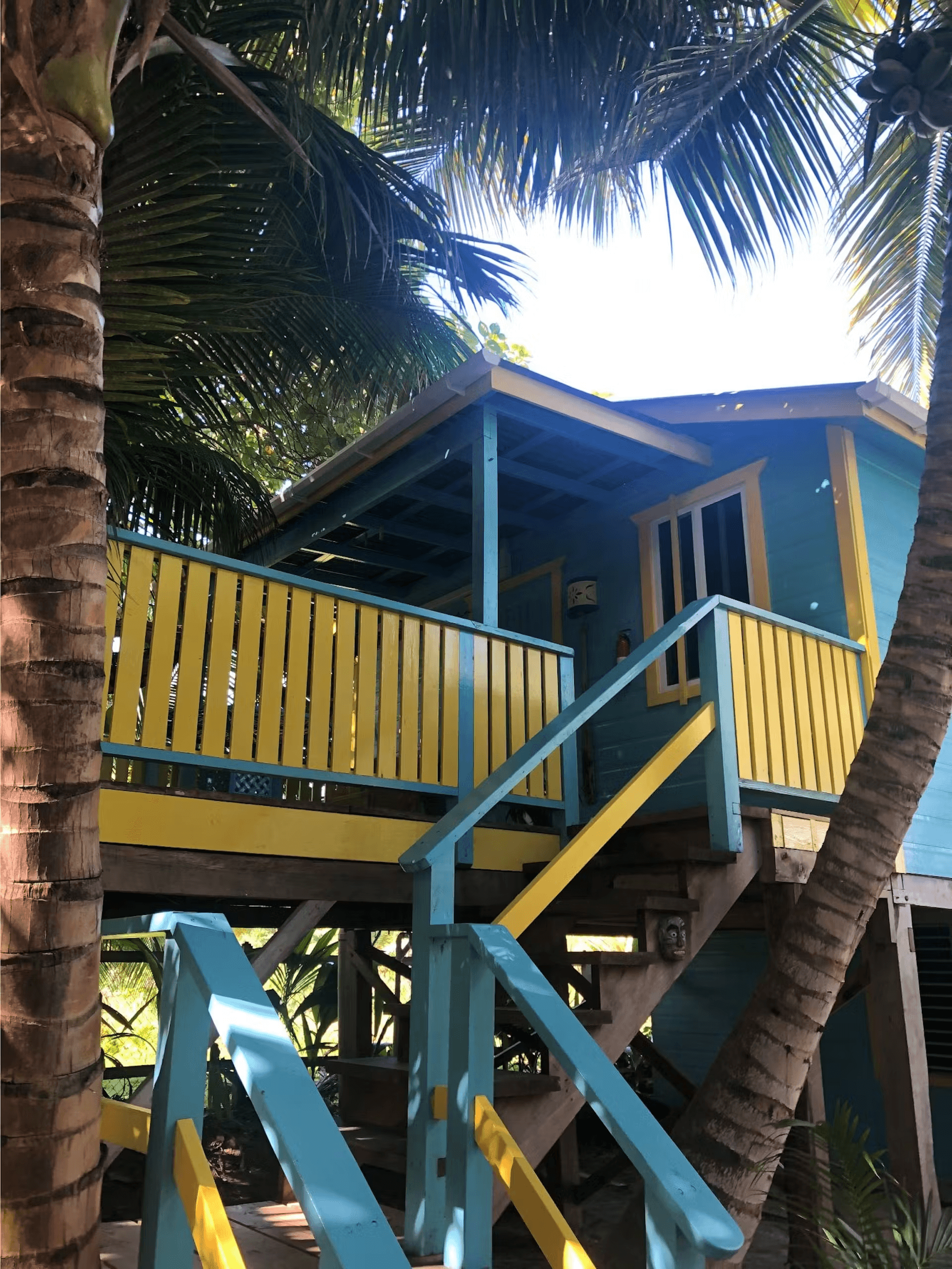 Exterior of a colorful blue and yellow painted cabana with stairs leading to second level, surrounded by palm trees