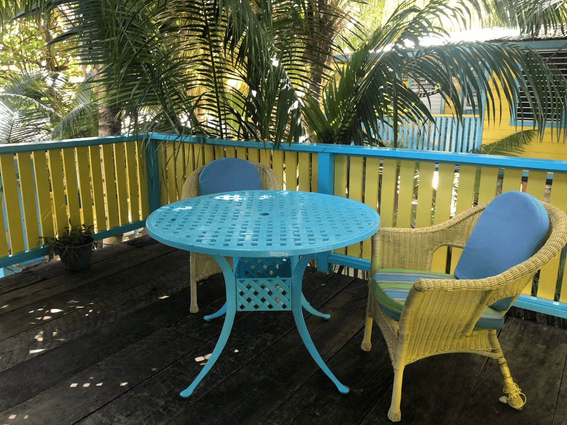 A balcony with yellow and blue rails and two yellow chairs with a round bright blue table, palm tree fronds form a backdrop