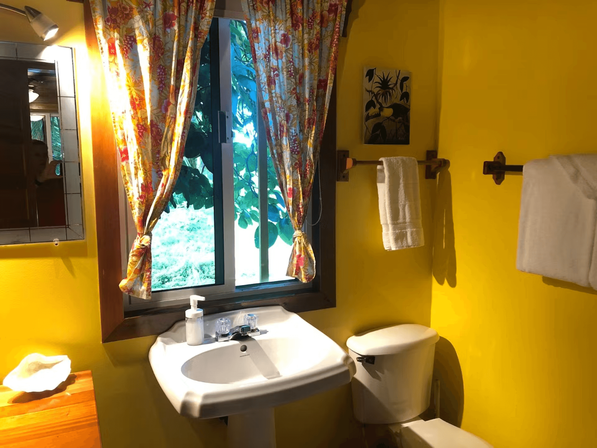 Bathroom painted bright yellow, a white pedestal sink with curtained window above, toilet, and white plush towels hanging on the wall