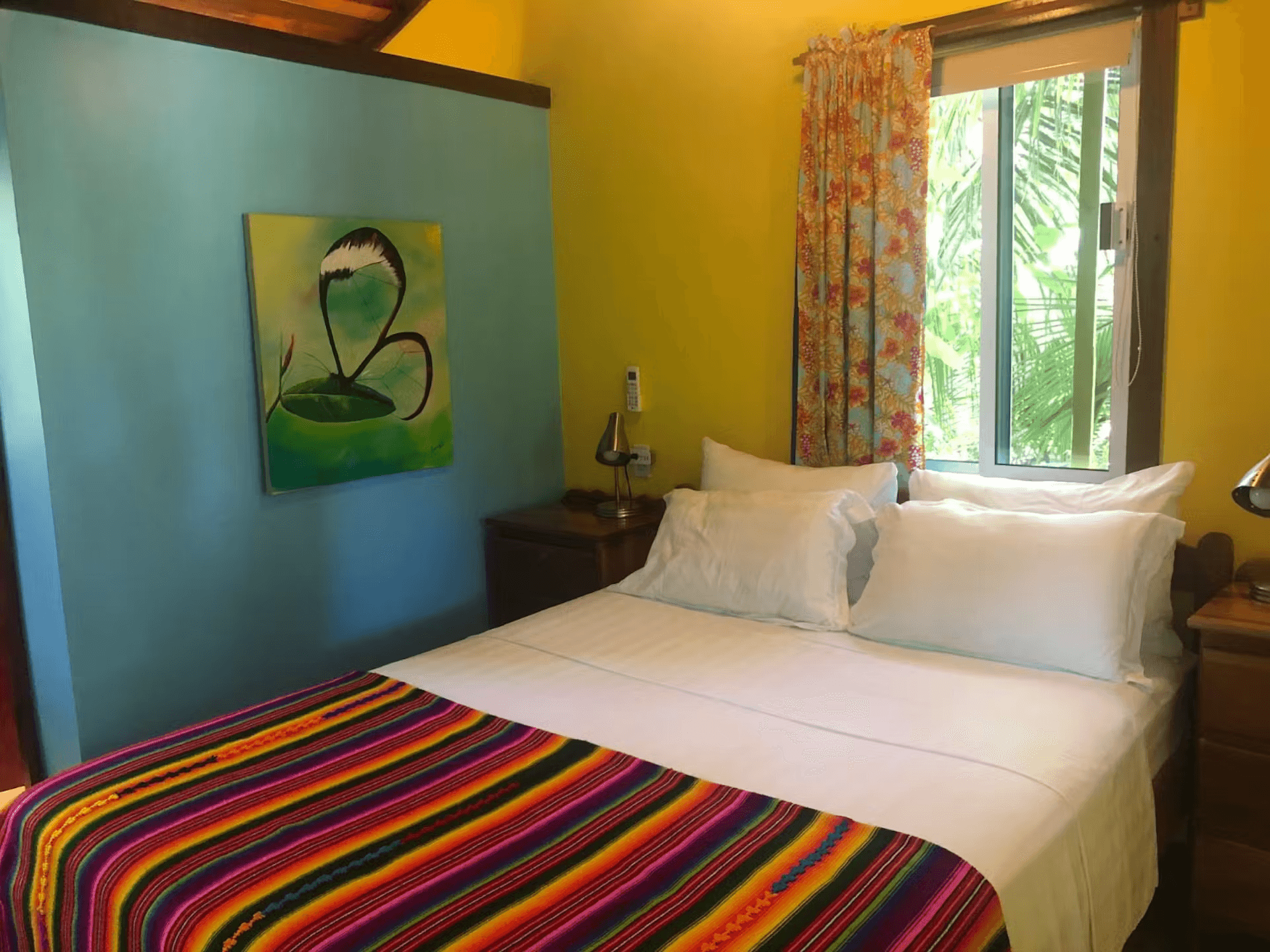 Blue and yellow painted bedroom with queen bed featuring striped bedspread and white sheets and pillowcases with sunny curtained window behind