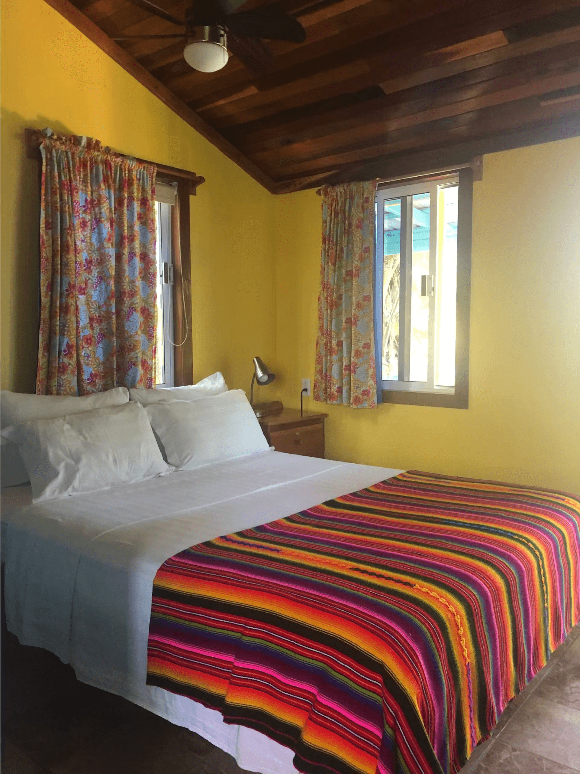 Bedroom with yellow painted walls, sun-filled windows with curtains, queen bed, and hardwood ceiling