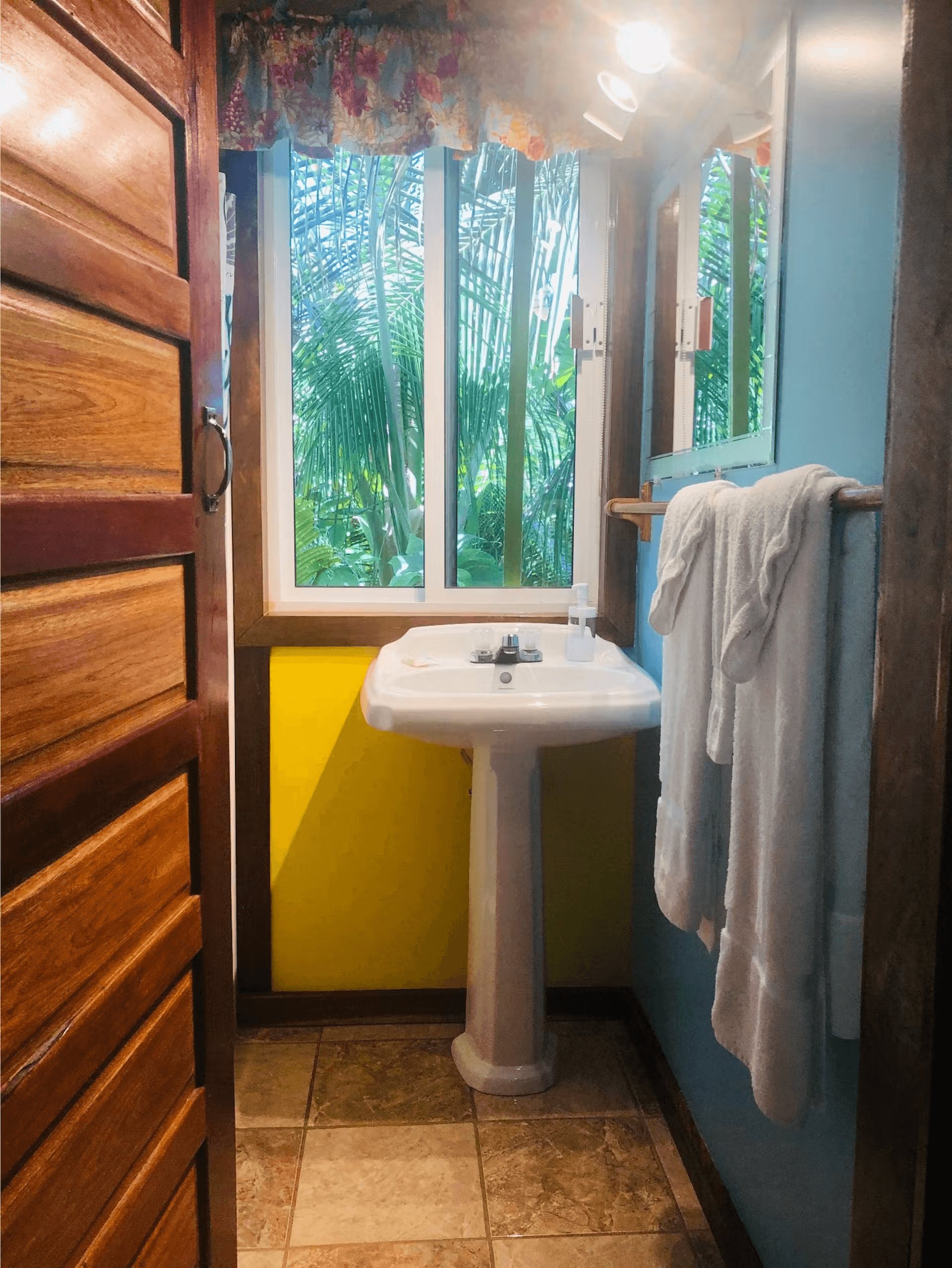 Bathroom with yellow and blue painted walls, white pedestal sink, with window above, and plush white towels hanging on wall