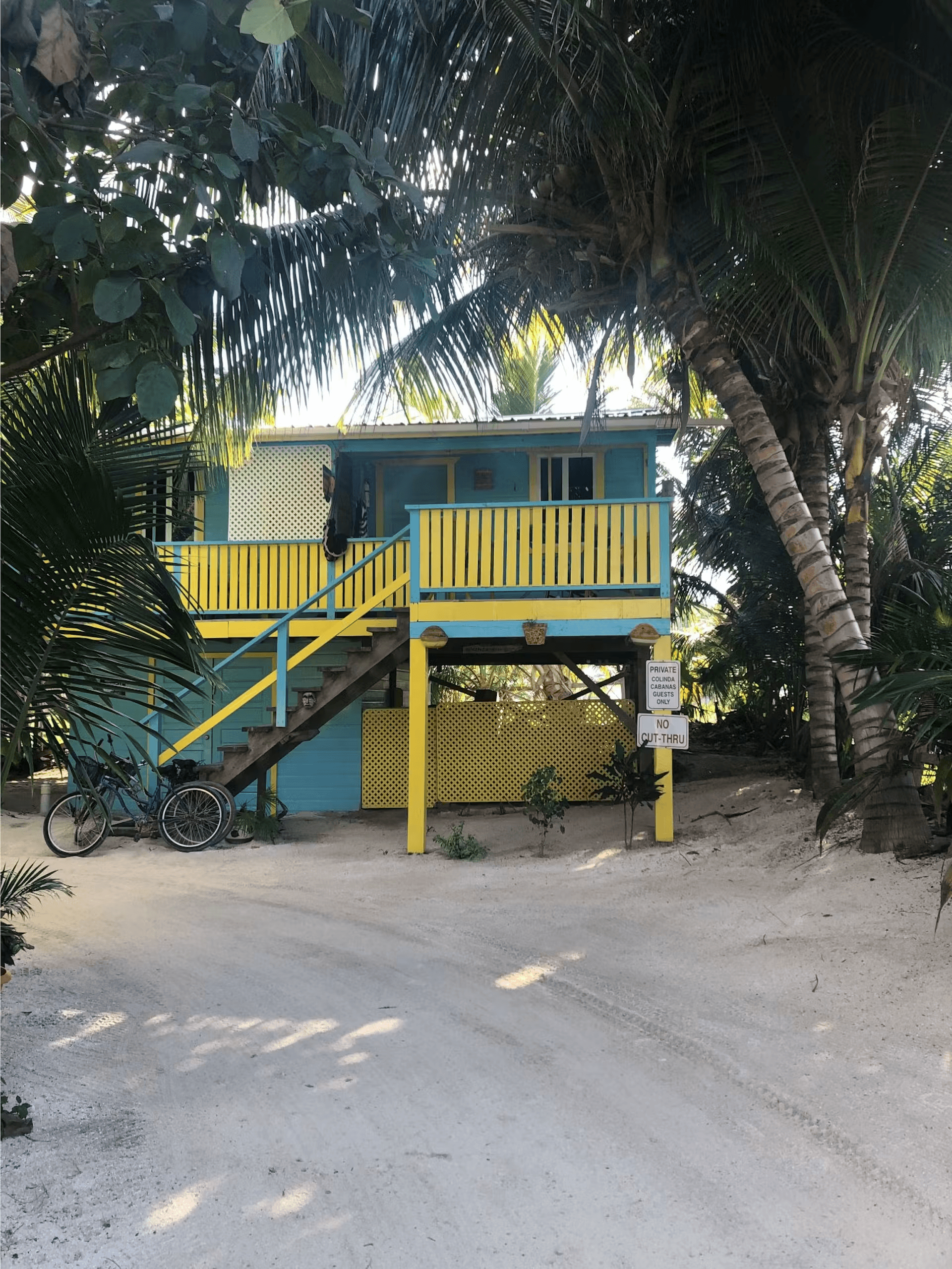 Exterior of brightly painted blue and yellow cabana with stairs leading to balcony, surrounded by sand and palm trees