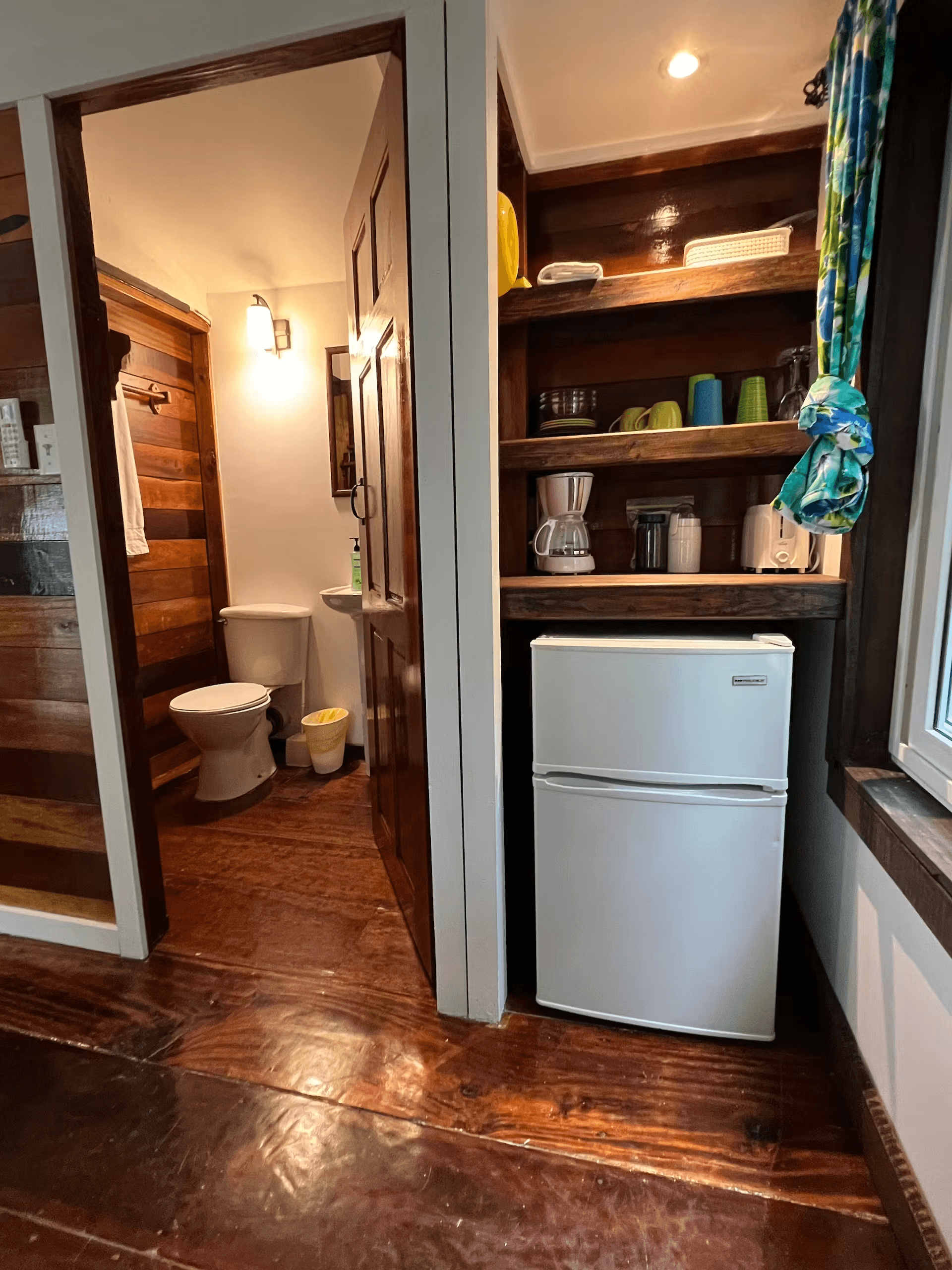 Built-in open shelves over small refrigerator next to window on one side and door to bathroom on the other