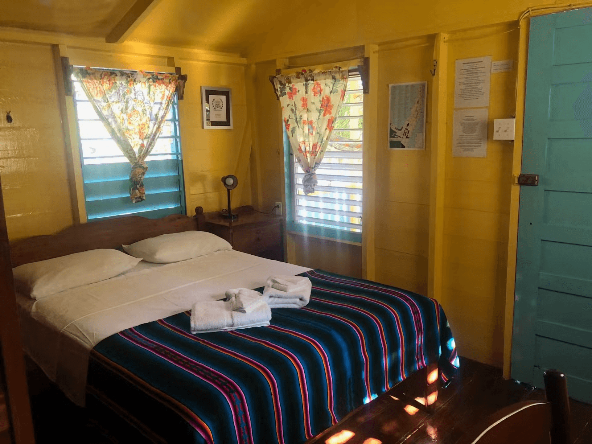 Bedroom with bright yellow walls, double bed with colorful striped blanket and white sheets, sunny windows with blinds and curtains