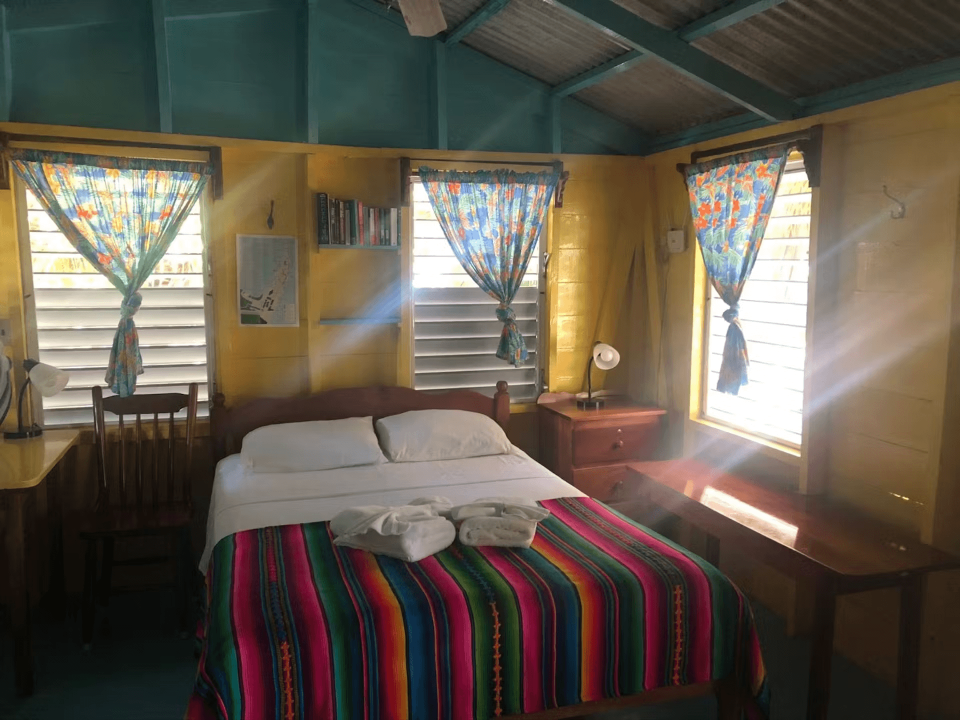 Sun streaming into bedroom with double bed, wood chair & nightstand with lamp, and vaulted painted wood ceiling