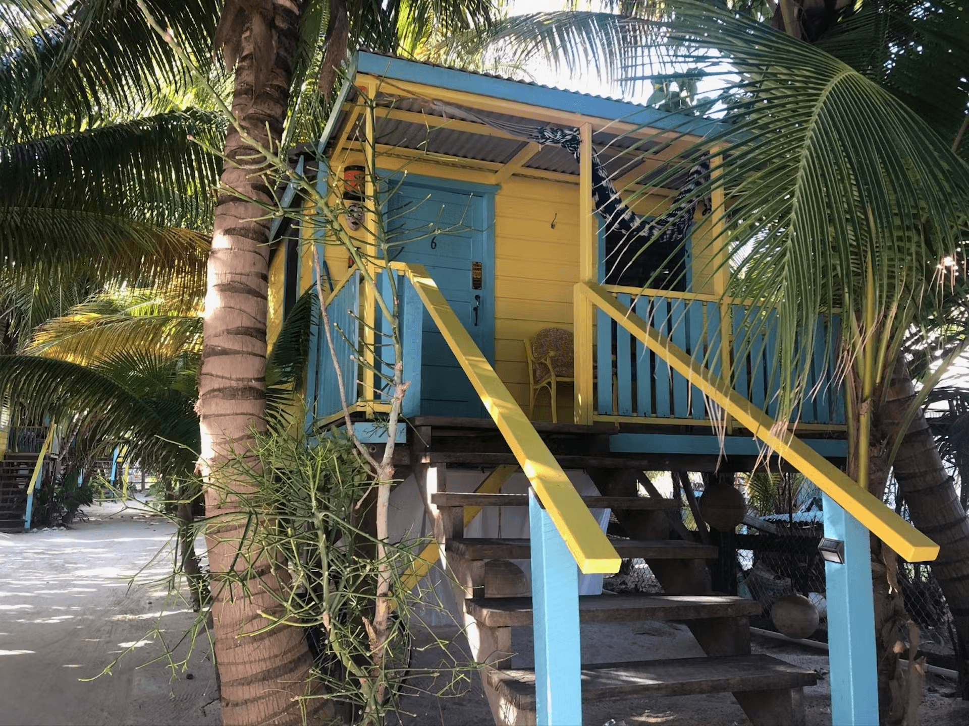 Exterior of a colorful blue and yellow cabana with stairs leading balcony and palm trees growing beside