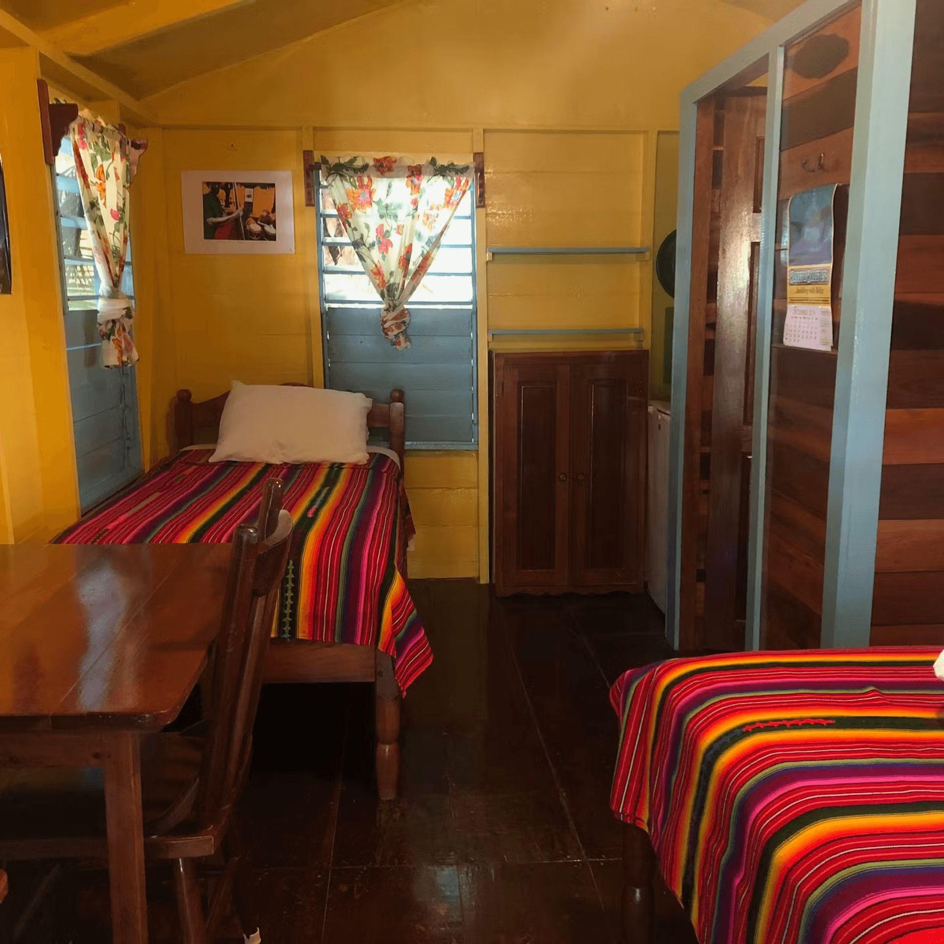 Bedroom showing foot of double bed, single bed on opposite wall and table with chair, sunny windows, and hardwood accents