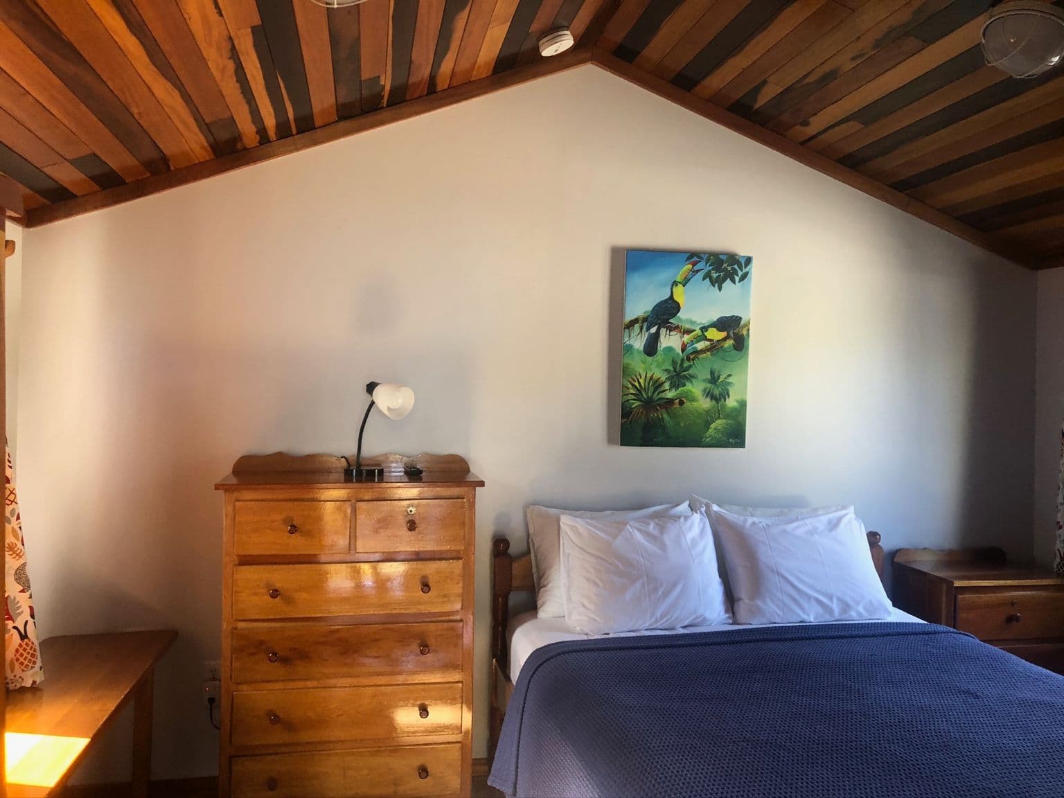 Bedroom with queen bed, wood dresser with table lamp, tropical artwork, and vaulted hardwood ceilings