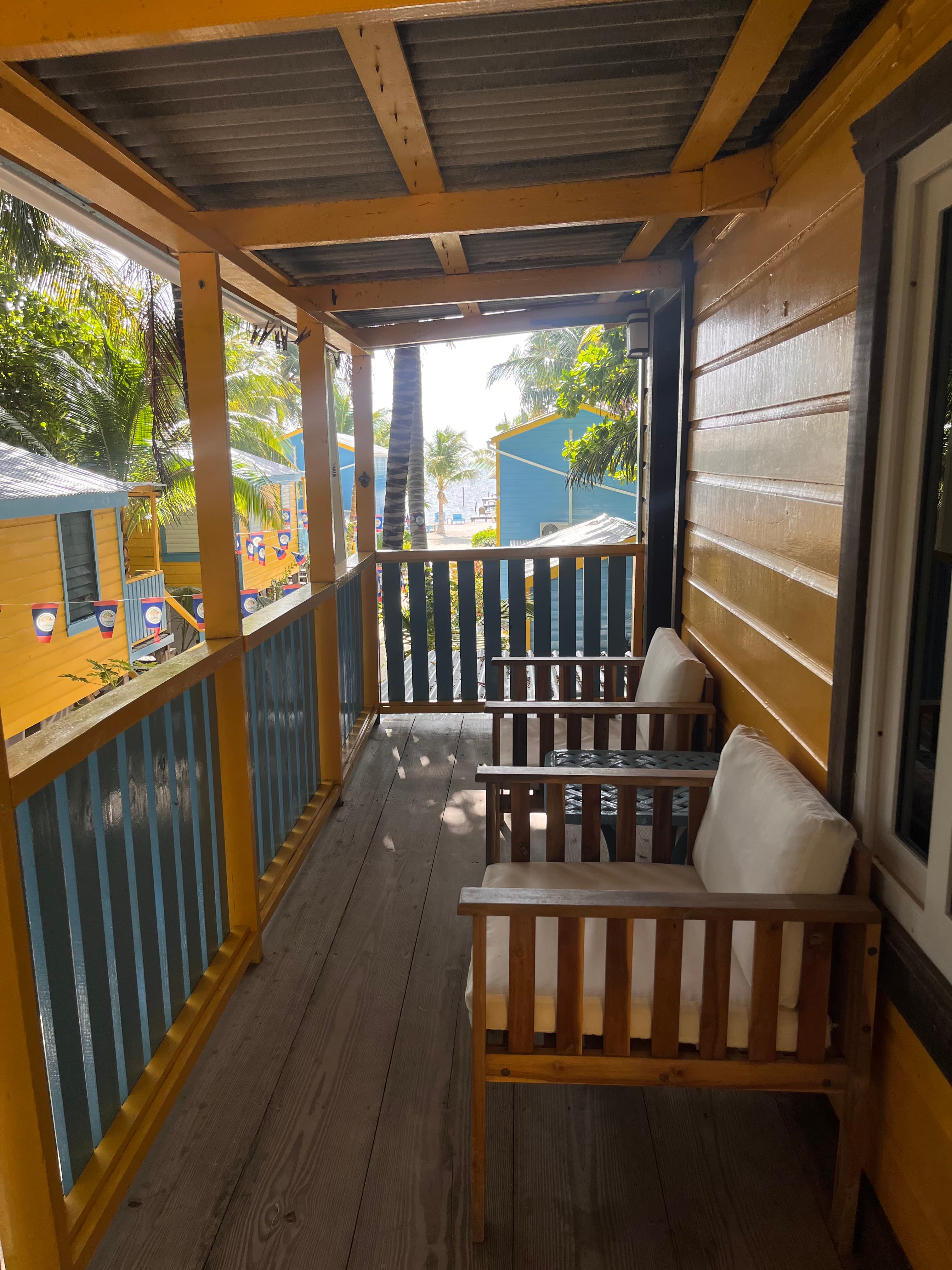 A wooden balcony with two white-cushioned chairs overlooks a tropical scene.