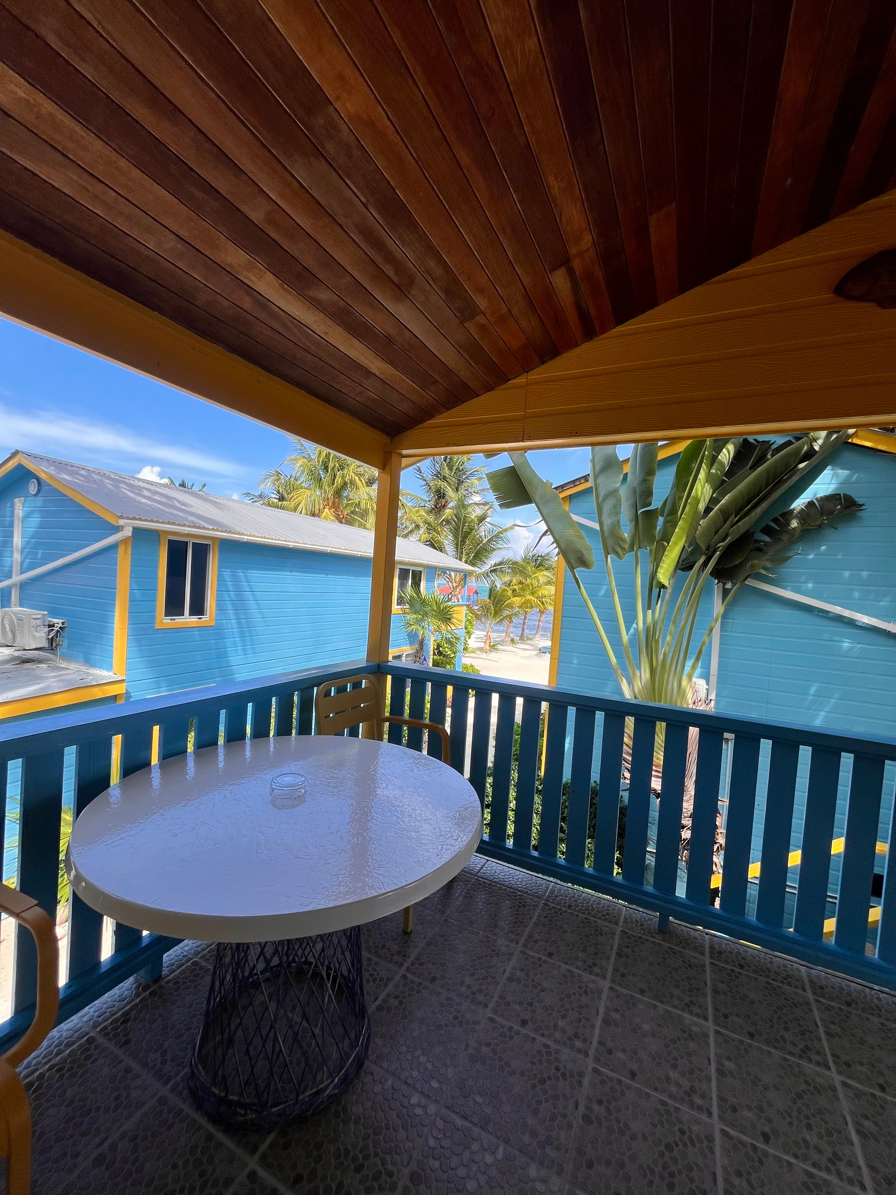 A cozy balcony with a white table and chair, surrounded by vibrant blue buildings and tropical greenery.