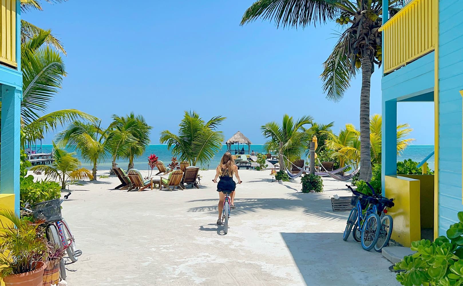 A person rides a scooter along a tropical path lined with palm trees and beach chairs, leading to the ocean in the background.