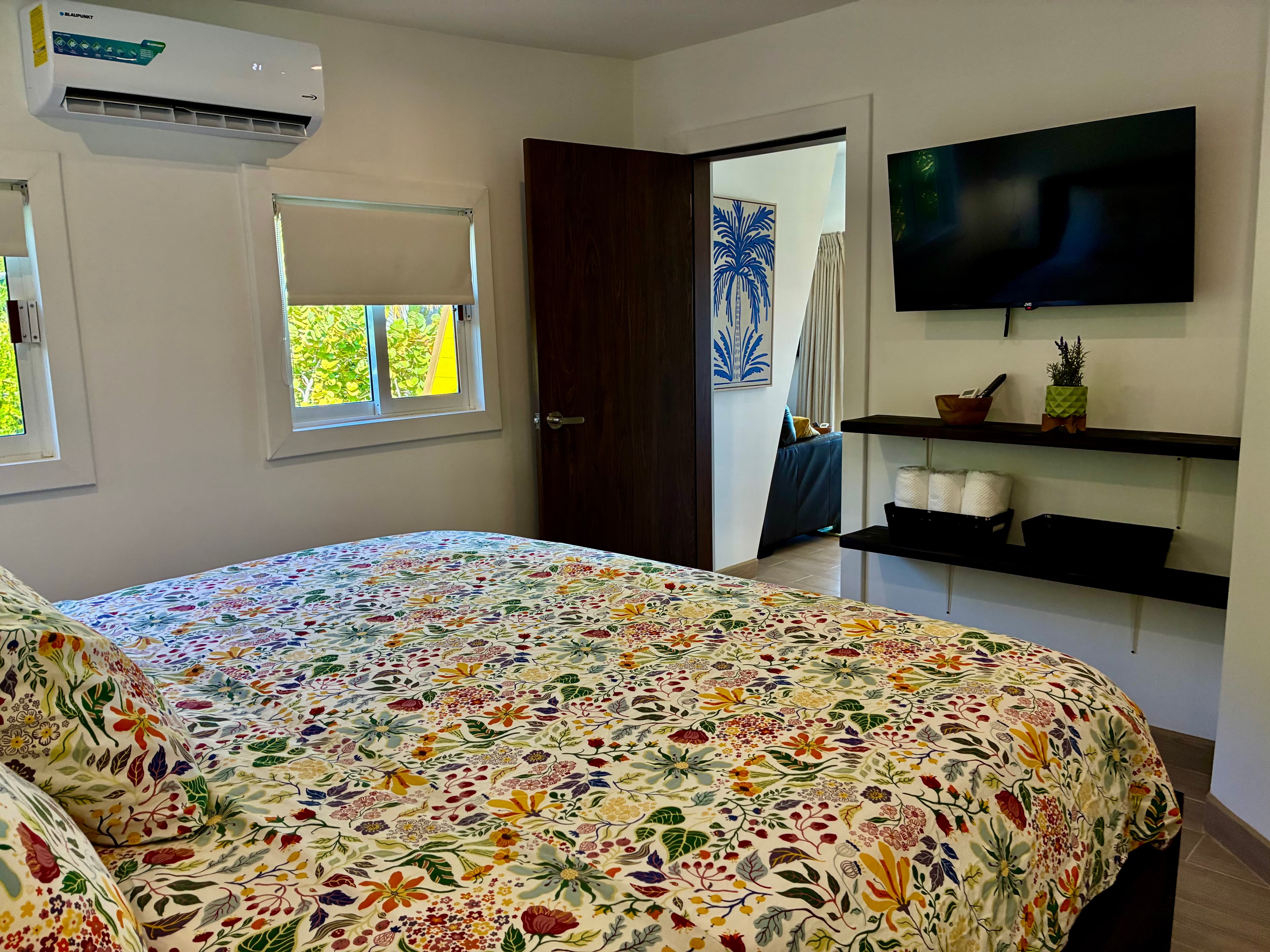A brightly decorated bedroom with a floral comforter, a television on the wall, and a door leading to another room.
