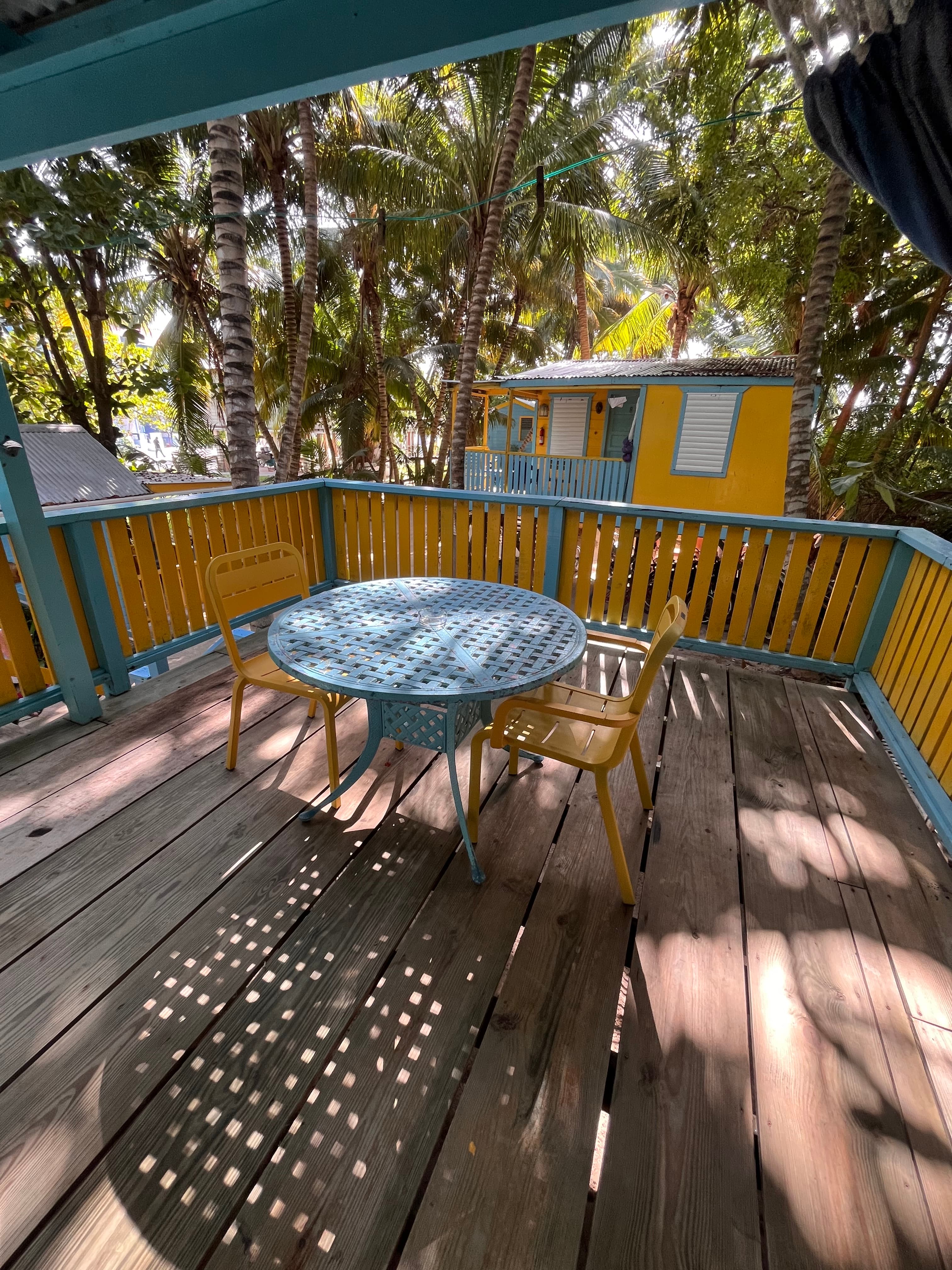 A bright turquoise and yellow deck features a round table with two chairs, surrounded by lush palm trees.
