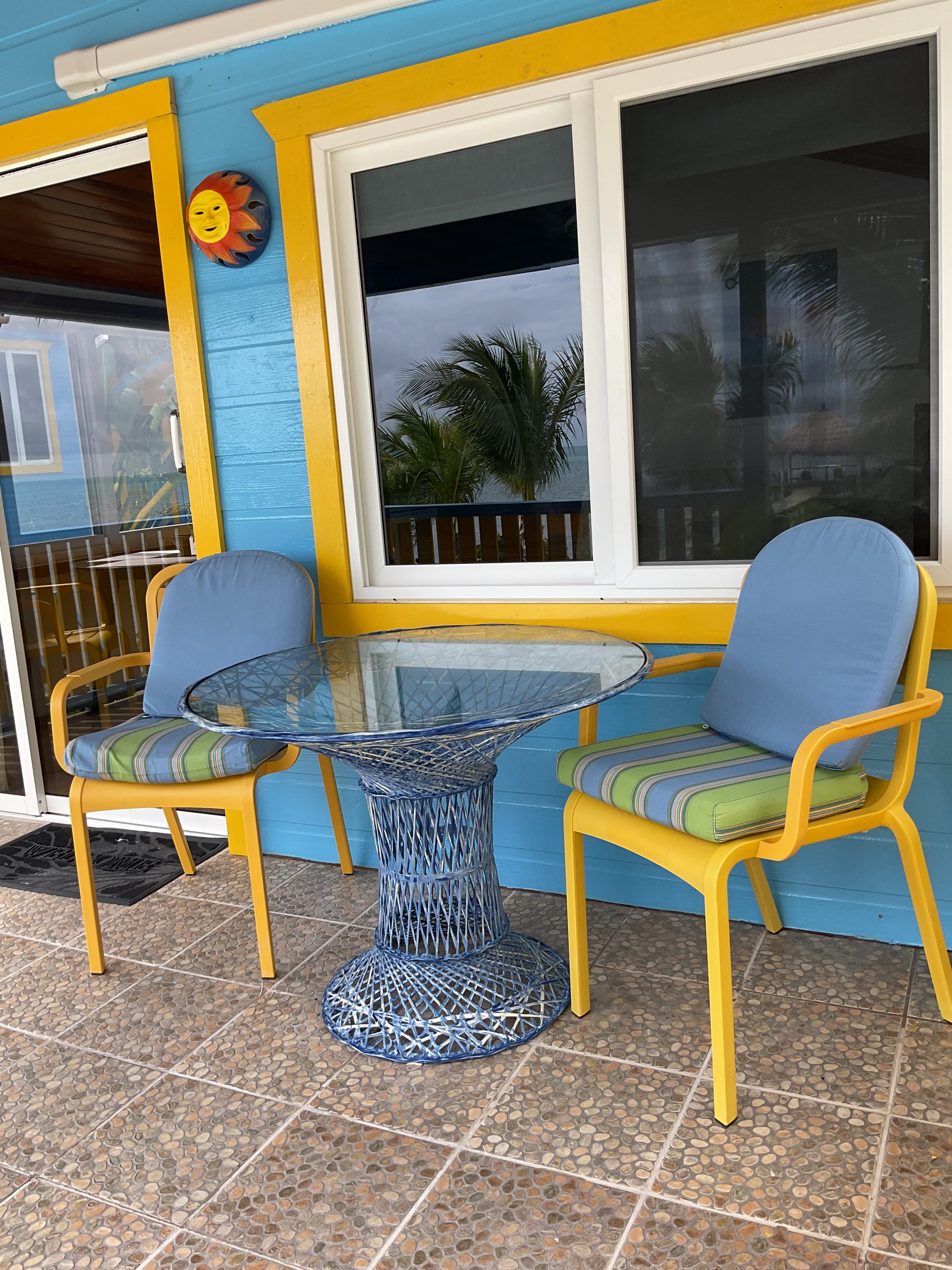 A glass-topped table with two colorful chairs on a porch with blue walls and a decorative sun.