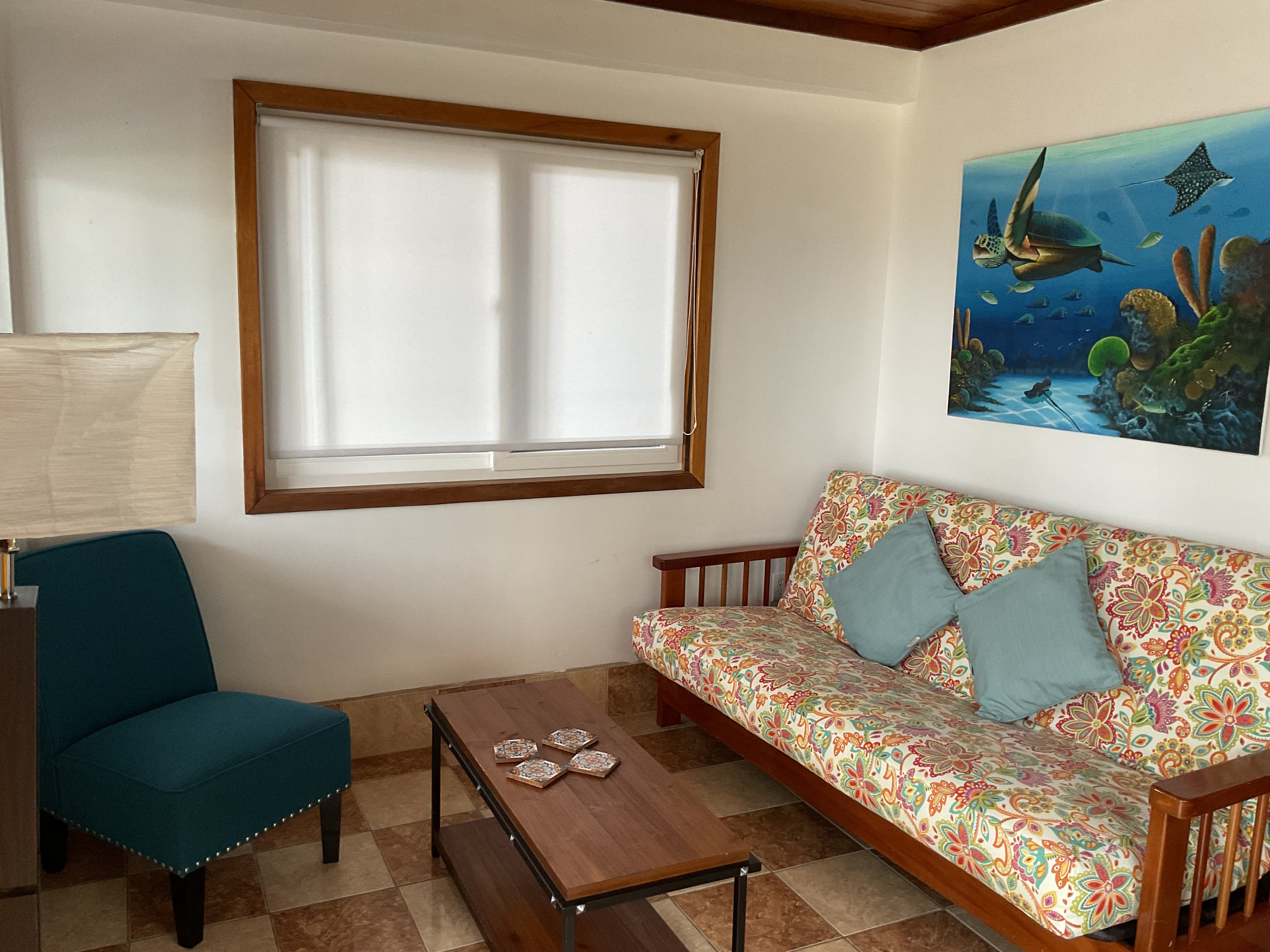 A cozy living room featuring a floral-patterned sofa, a turquoise armchair, a wooden coffee table, and ocean-themed wall art.