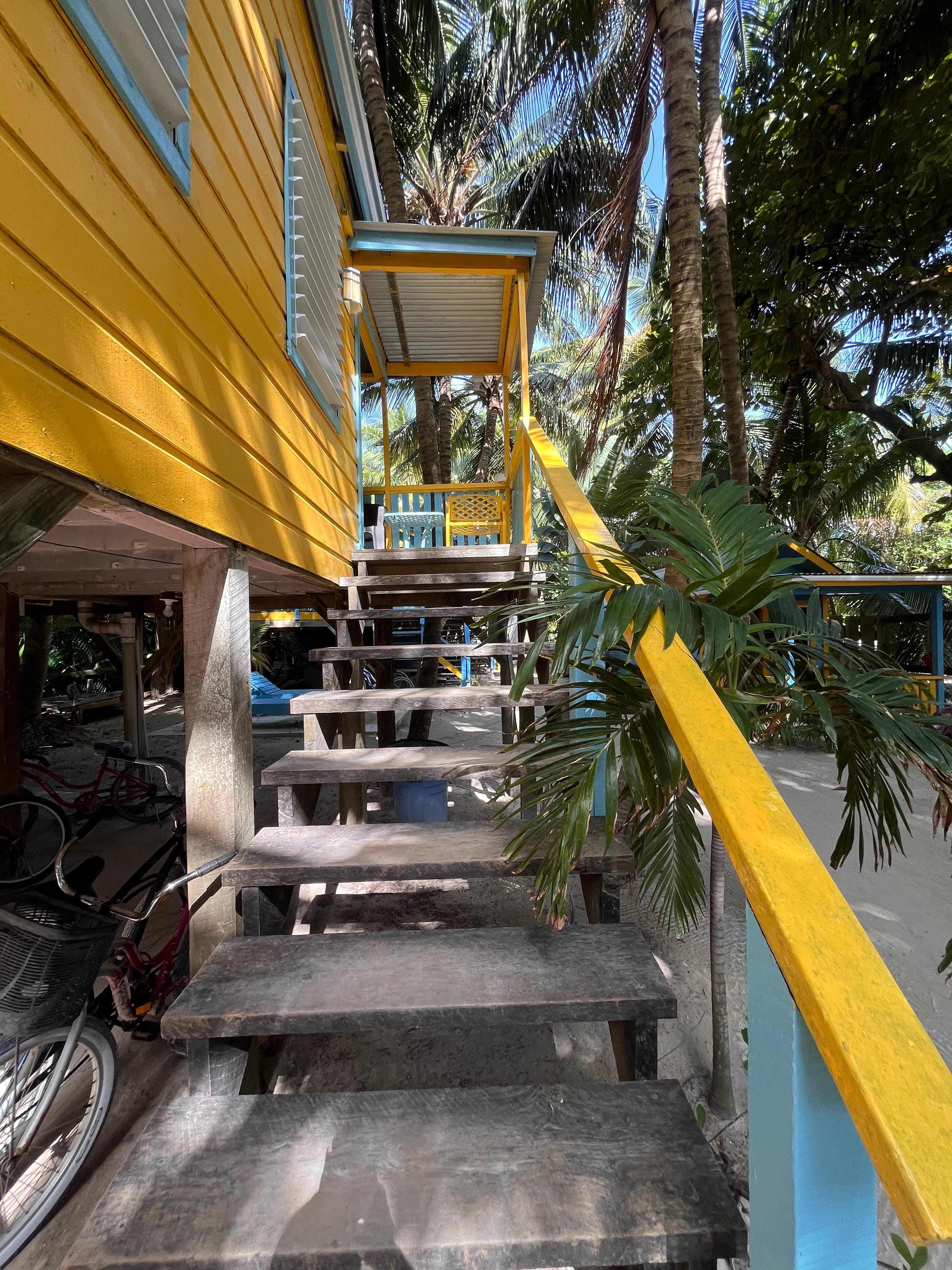 A set of wooden stairs leads up to a vibrant yellow building surrounded by tropical greenery.