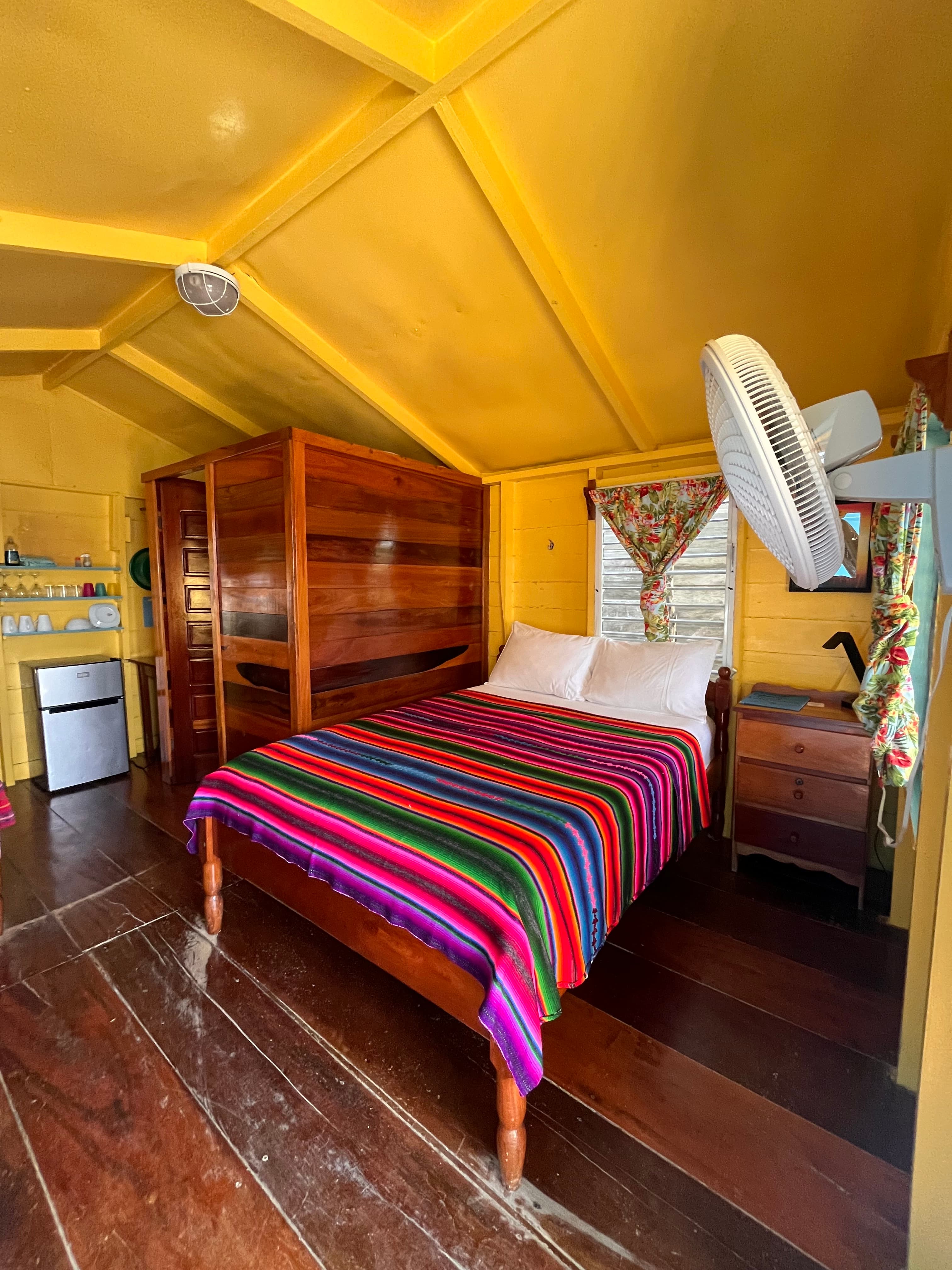 A cozy bedroom with a colorful striped blanket, wooden furniture, and bright yellow walls.