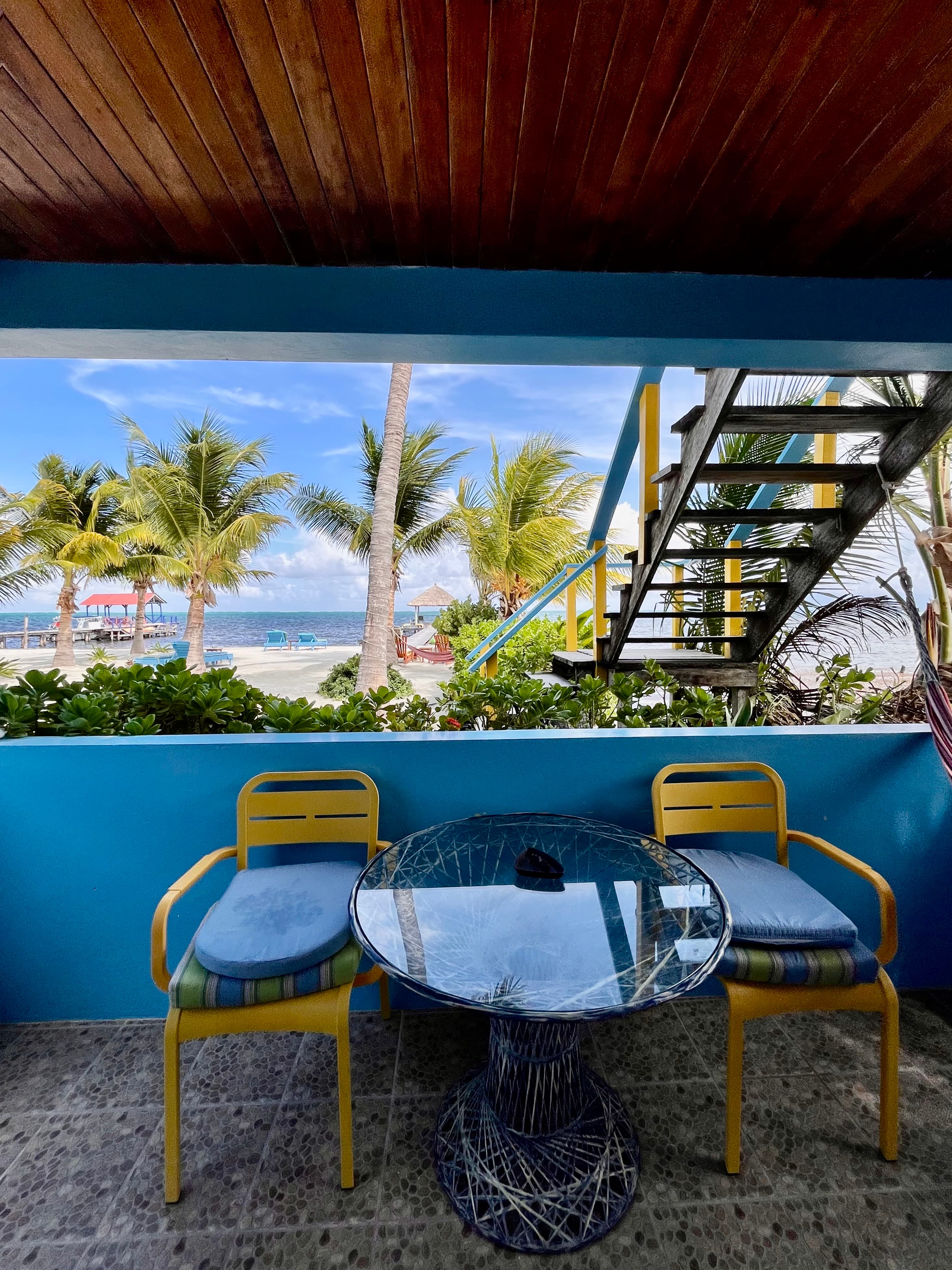 Tropical view from a balcony featuring a glass table and colorful chairs, overlooking a beach with palm trees.