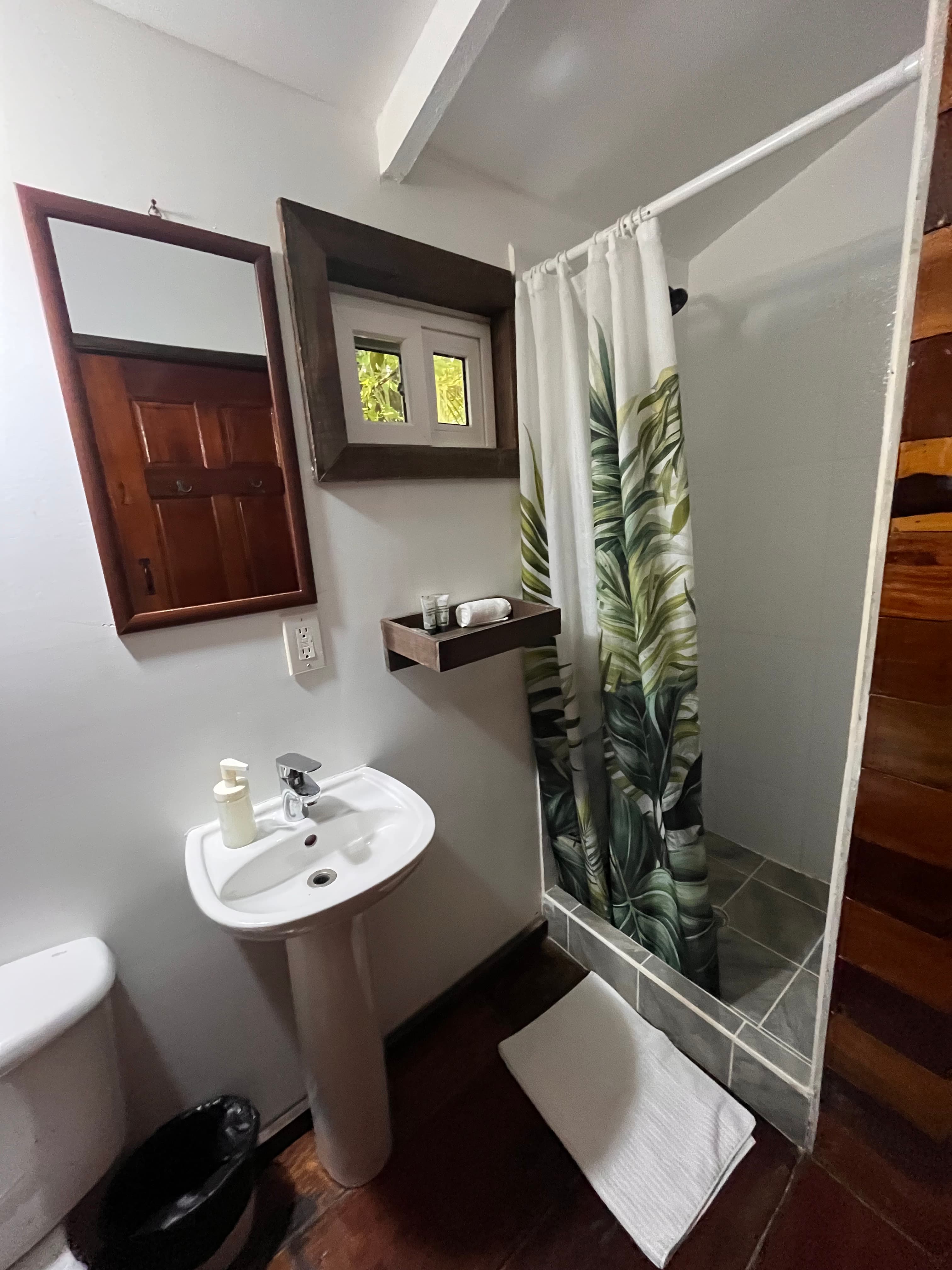A modern bathroom featuring a shower with a tropical-themed curtain, a pedestal sink, and a small shelf with toiletries.