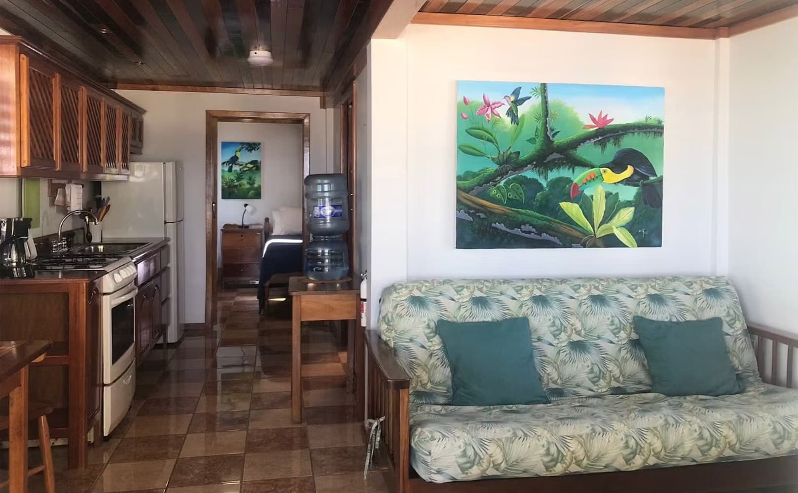 A bright living space featuring a kitchen, water cooler, and a couch adorned with tropical cushions, complemented by colorful wall art.