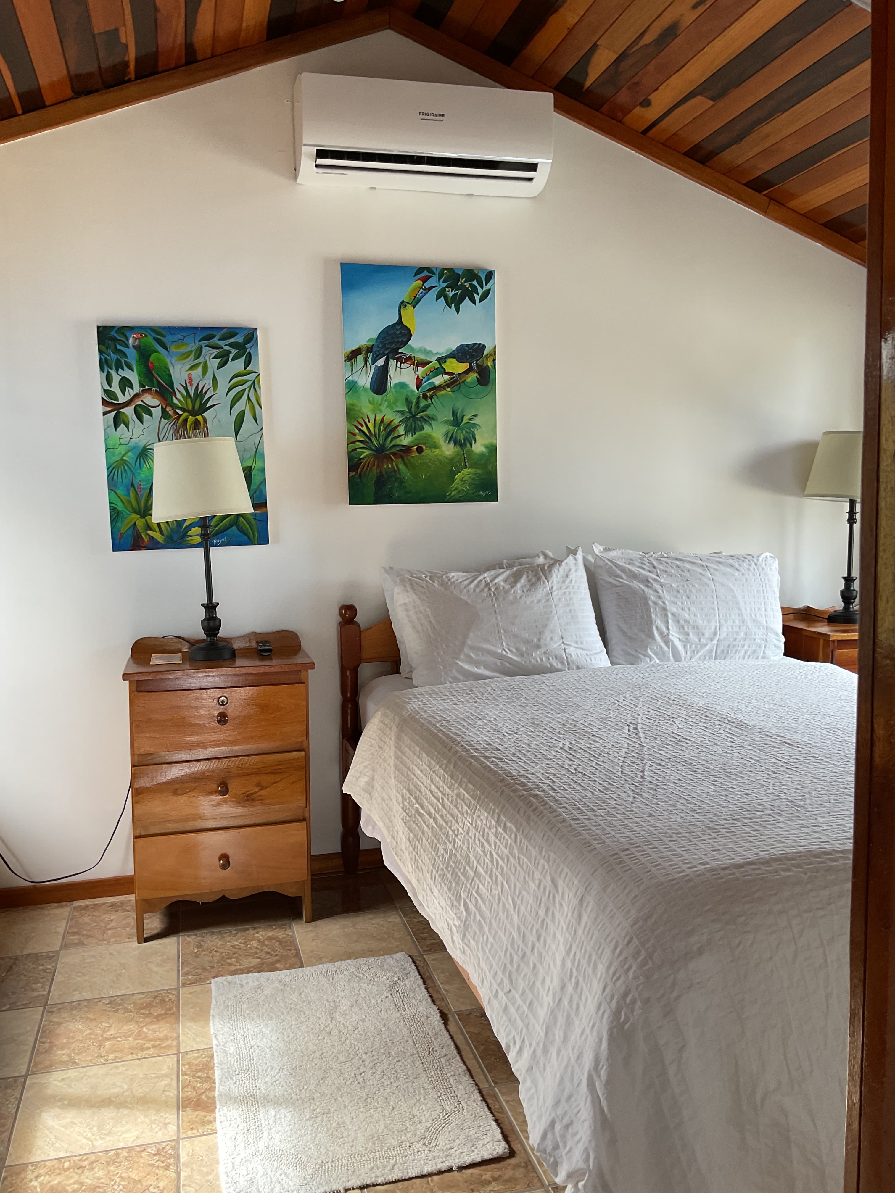 A cozy bedroom featuring a wooden bed, two art pieces of tropical scenery, and a bedside lamp.