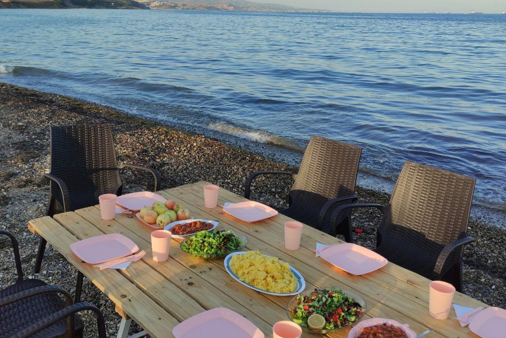 A wooden table set with plates of food and drinks, surrounded by chairs, beside a calm beach.