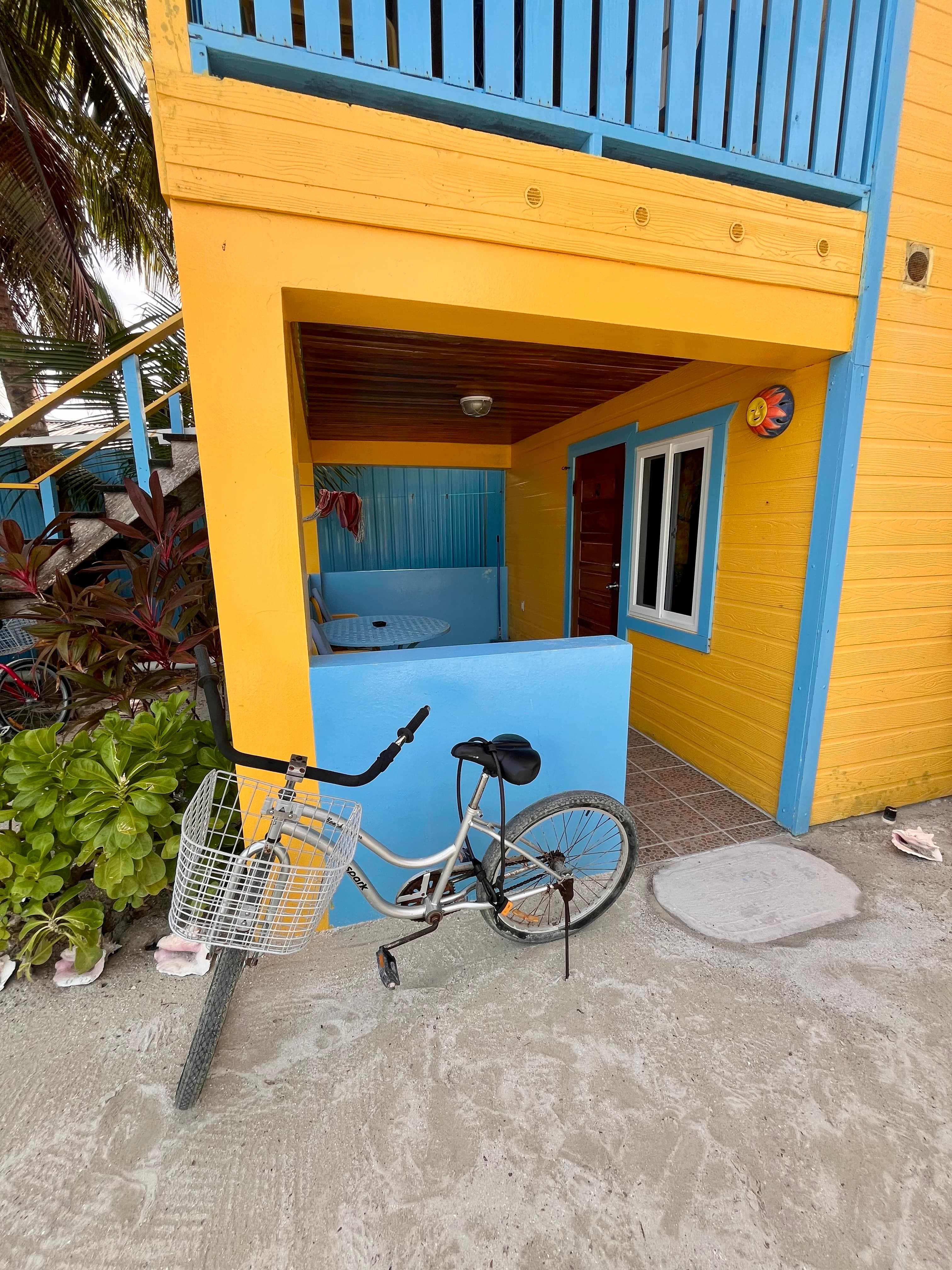 A white bicycle is parked beside a vibrant yellow and blue building with a porch.