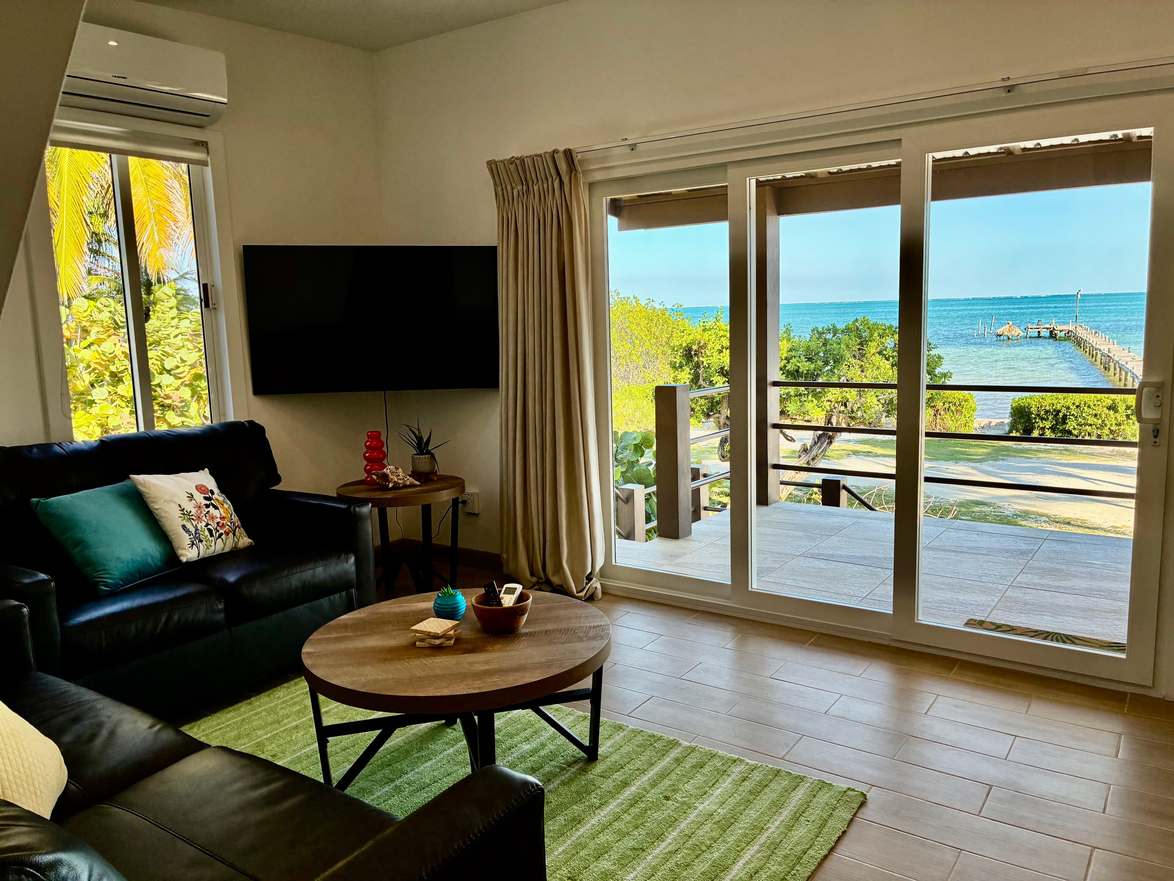 A cozy living room with a view of the beach, featuring a black sofa, coffee table, and large glass doors.
