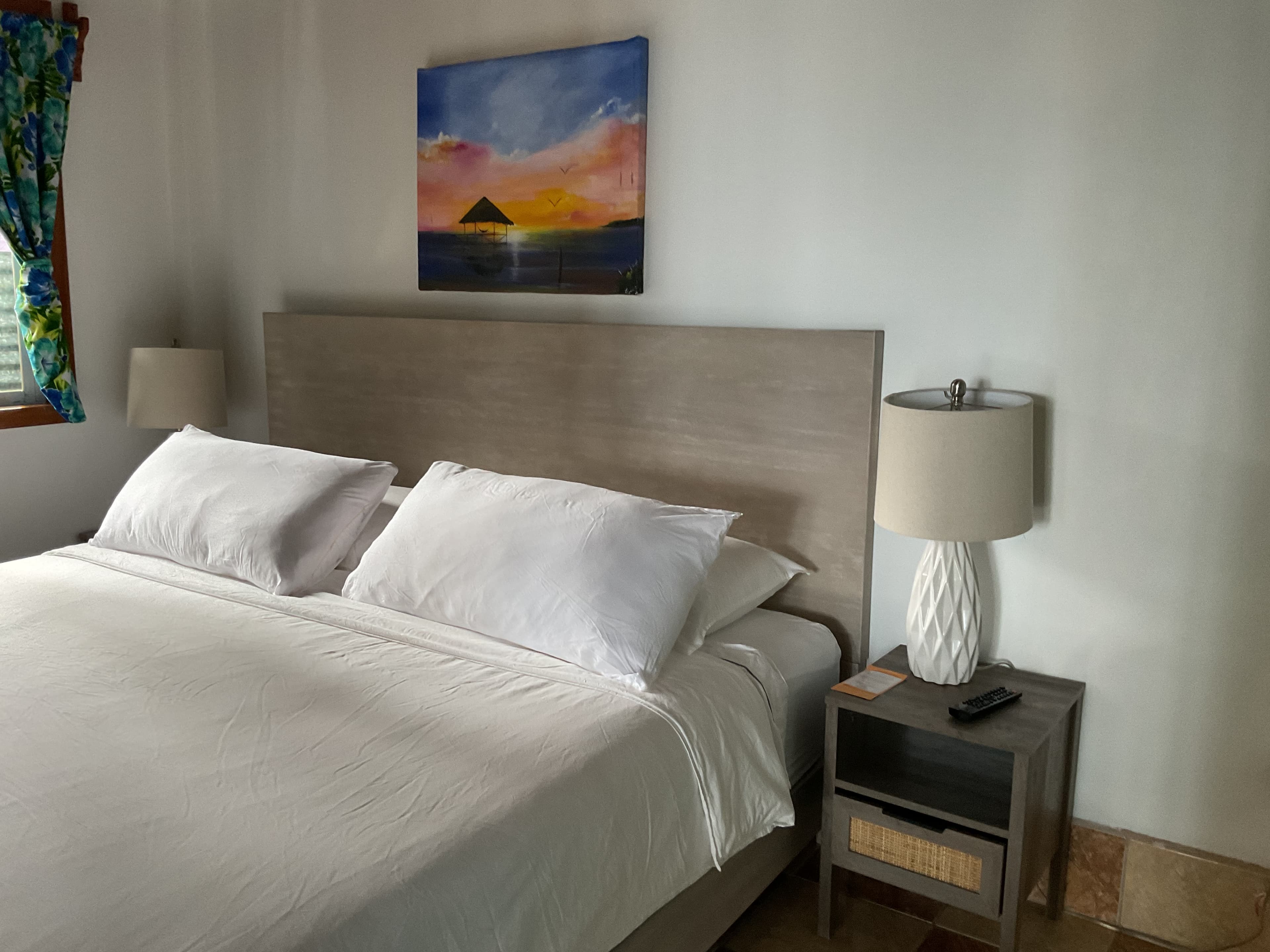 A neatly made bed with white linens and pillows, accompanied by a bedside table with a lamp and remote, and a colorful painting on the wall.
