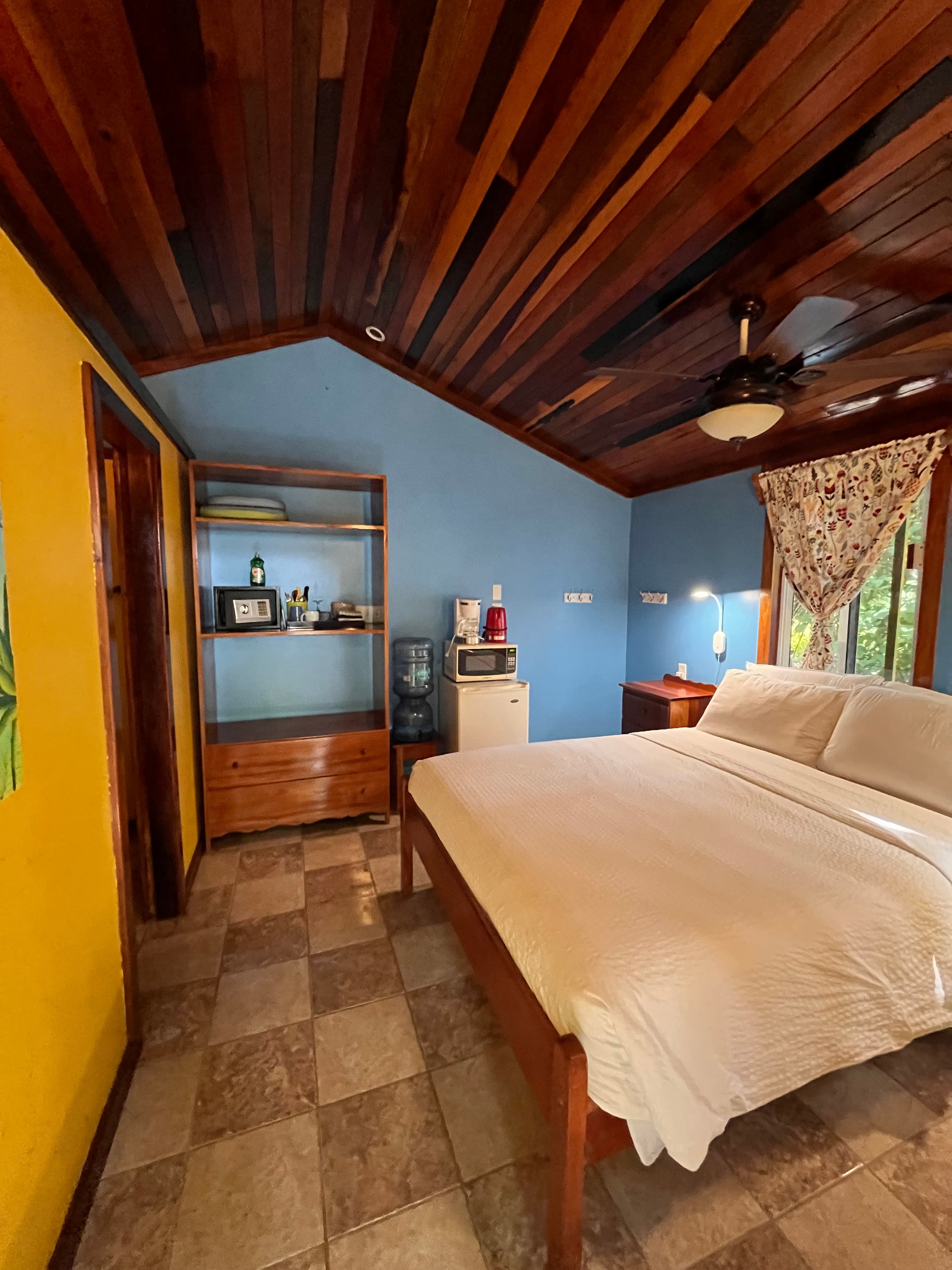 A cozy bedroom featuring a wooden ceiling, a queen bed, and a small kitchenette area with a shelf.