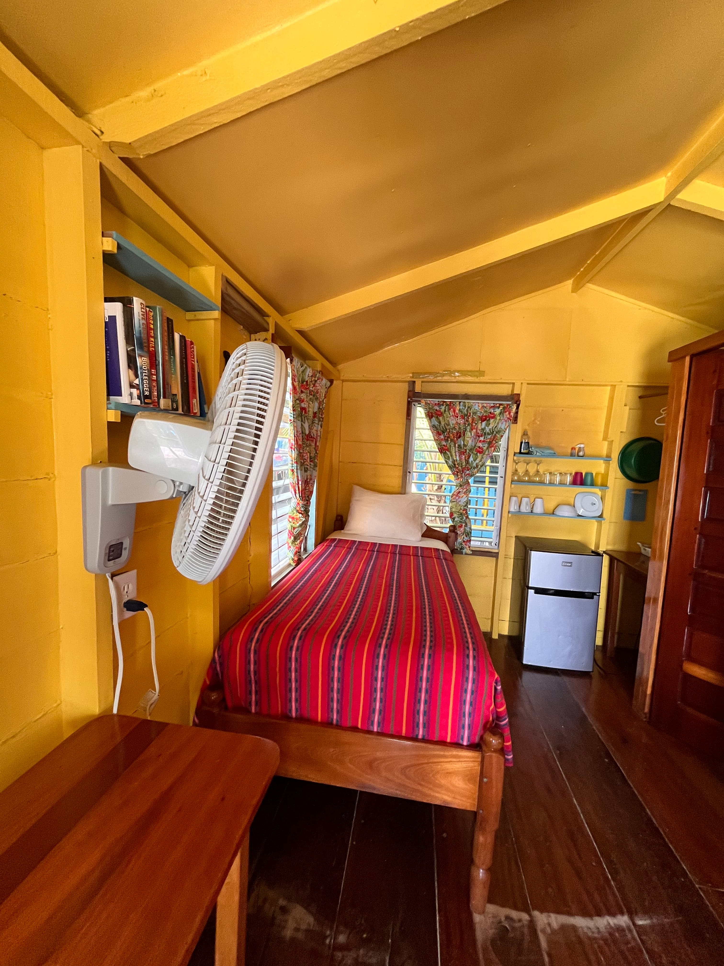 A cozy yellow-walled cabin featuring a single bed with a striped red blanket, a fan, bookshelves, and a small refrigerator.