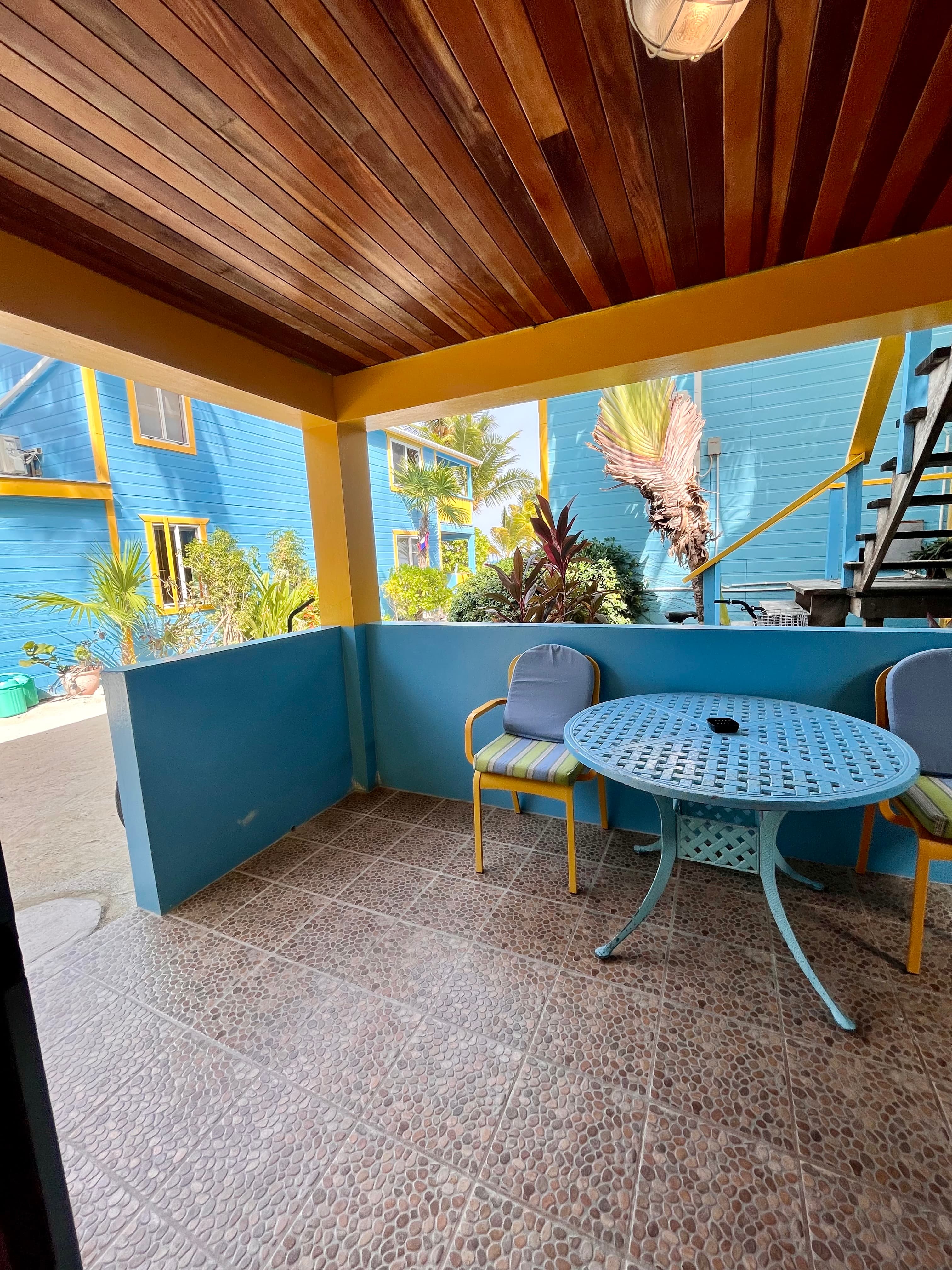 A cozy outdoor seating area with a blue table and chairs, surrounded by tropical plants and colorful buildings.