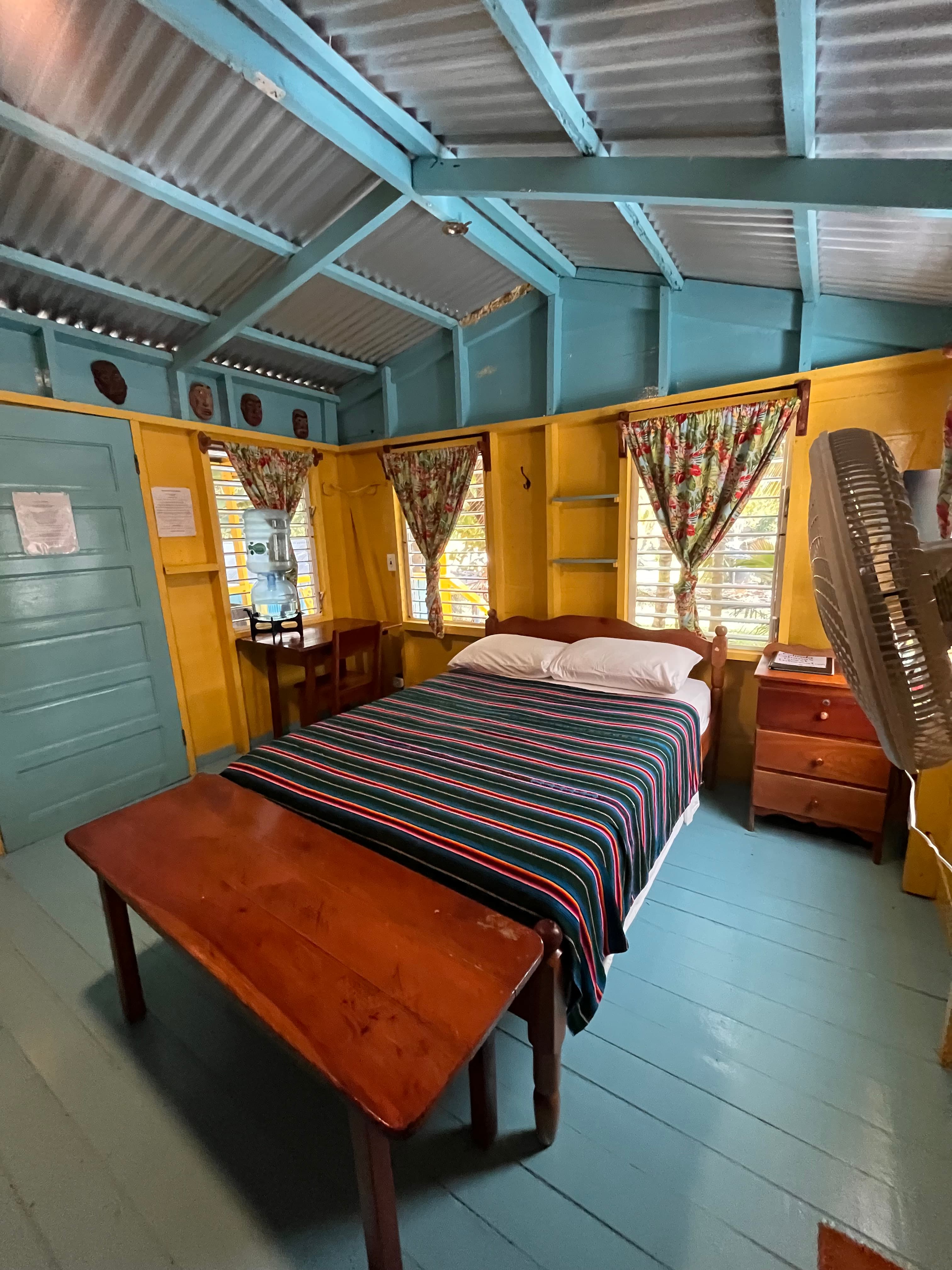 A brightly colored bedroom featuring a striped bedspread, wooden furniture, and large windows with floral curtains.