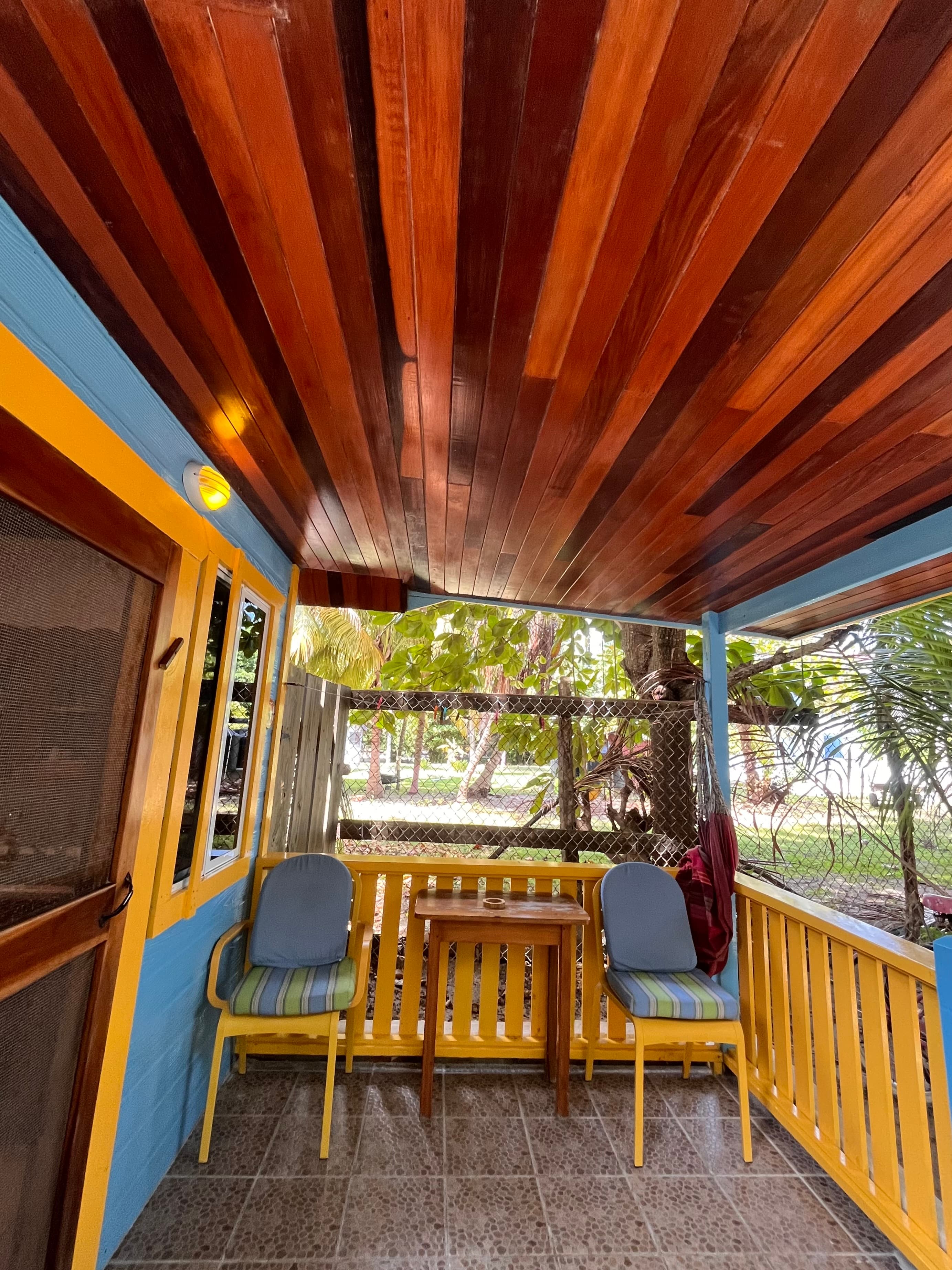A cozy porch featuring wooden ceiling beams, yellow and blue accents, and two chairs beside a small table, overlooking a green yard.