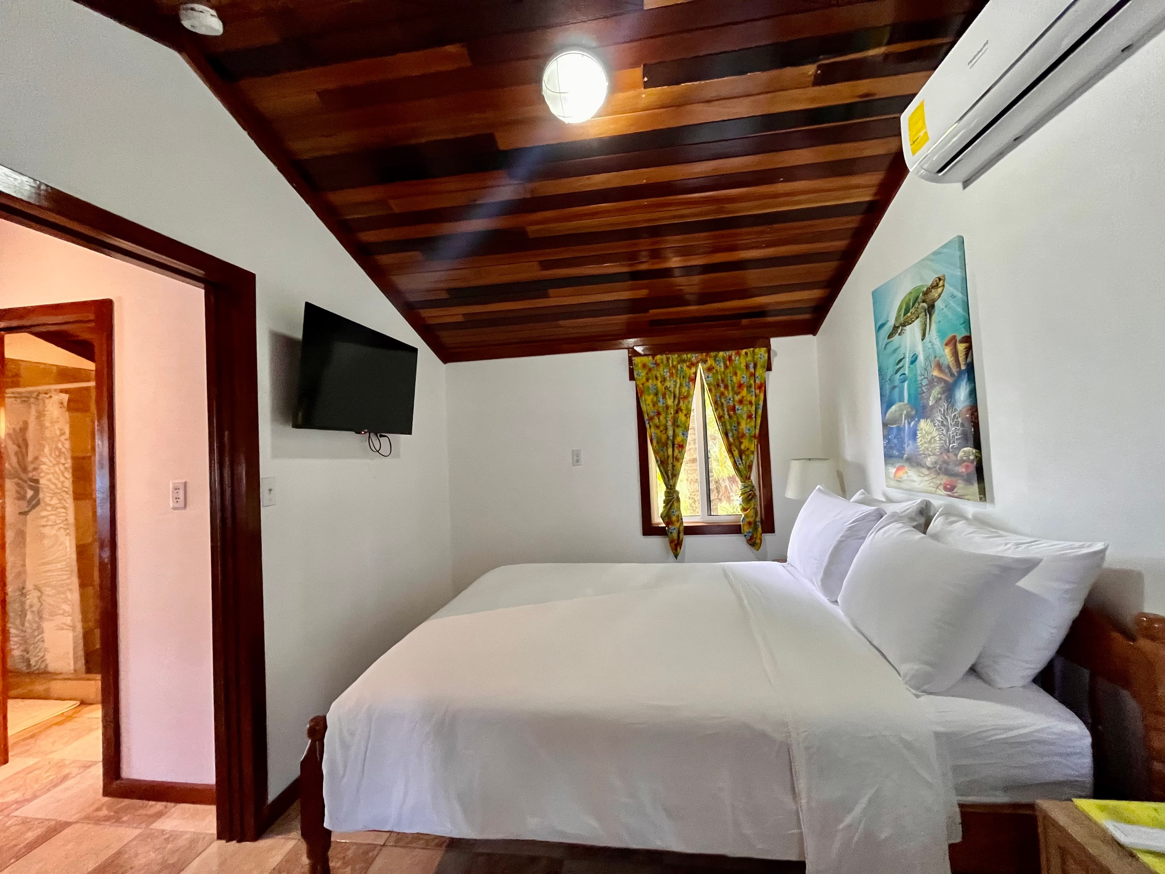 A cozy bedroom featuring two beds, a wooden ceiling, a television, and a window with colorful curtains.
