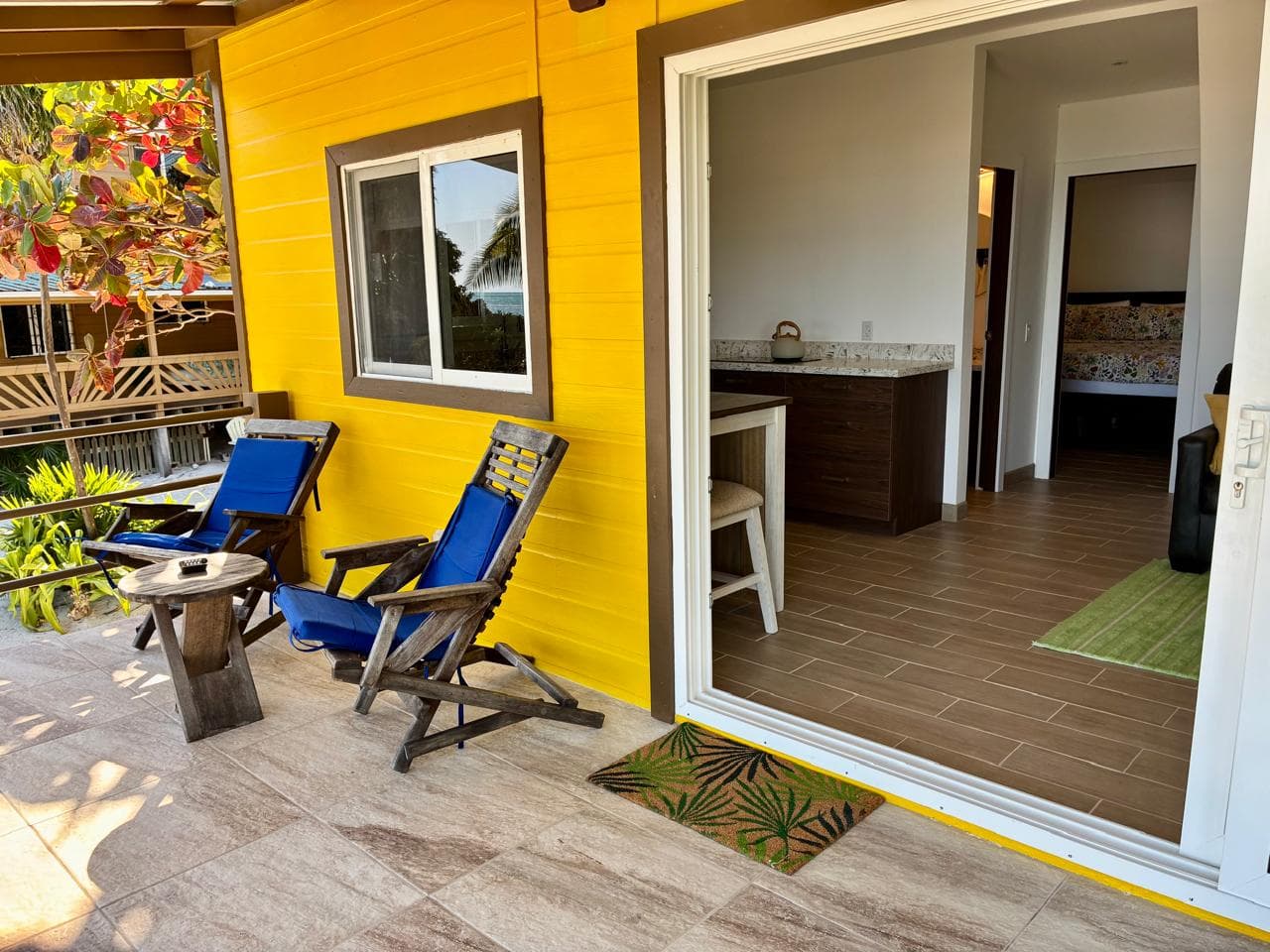 A bright yellow exterior with two blue lounge chairs on a patio leads to an indoor kitchen area.