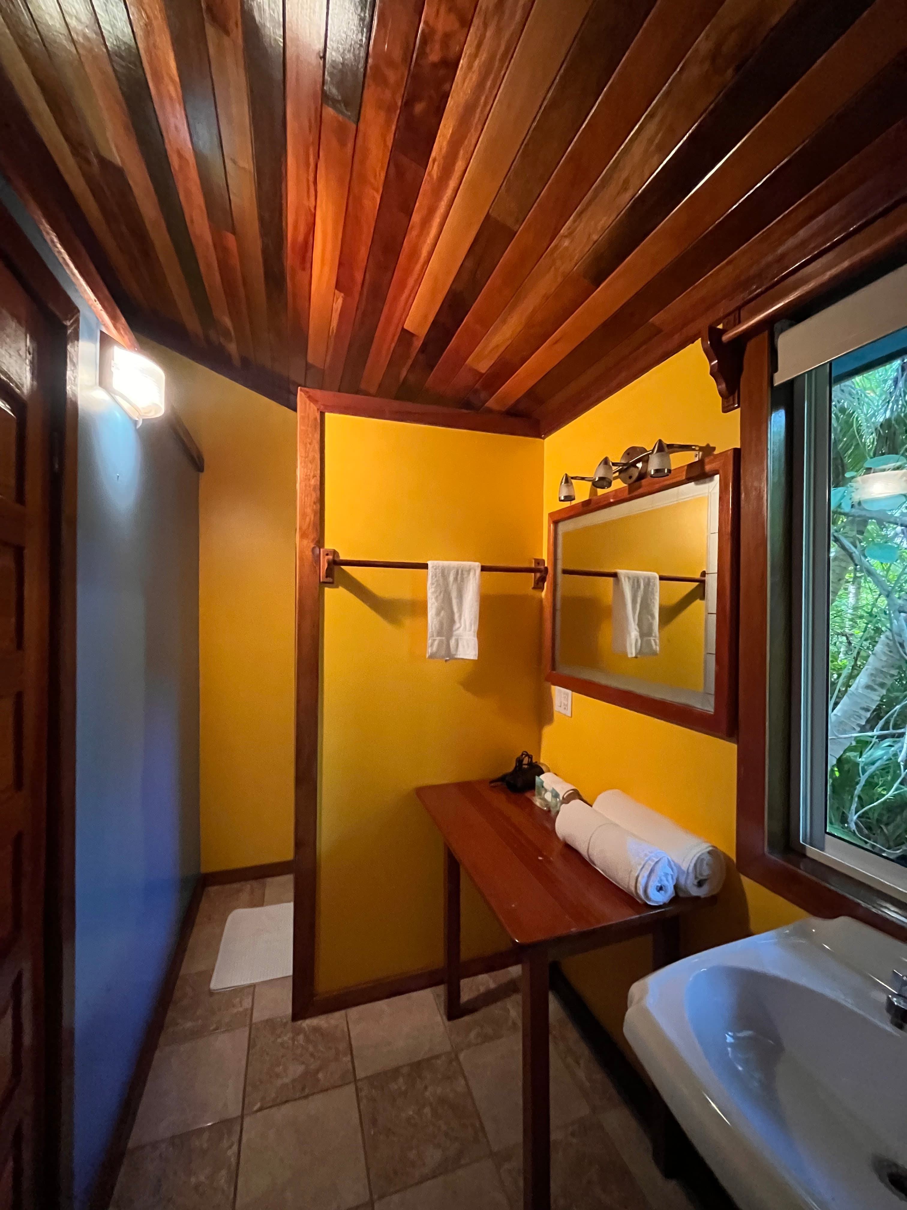 Brightly lit bathroom featuring wooden accents, yellow walls, a wooden table with rolled towels, and a mirror.