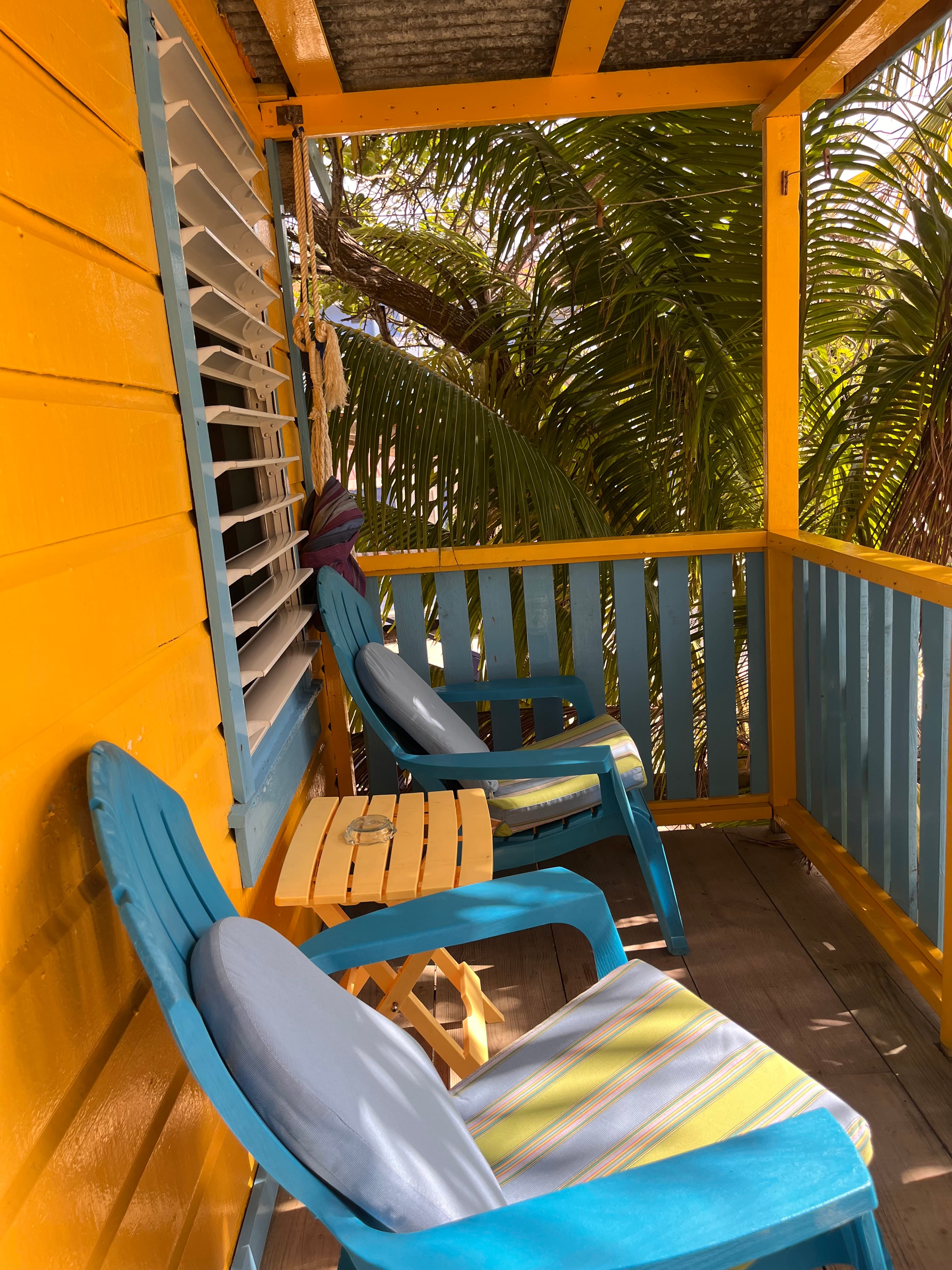 A sunny porch featuring two colorful chairs, a small table, and lush palm greenery in the background.
