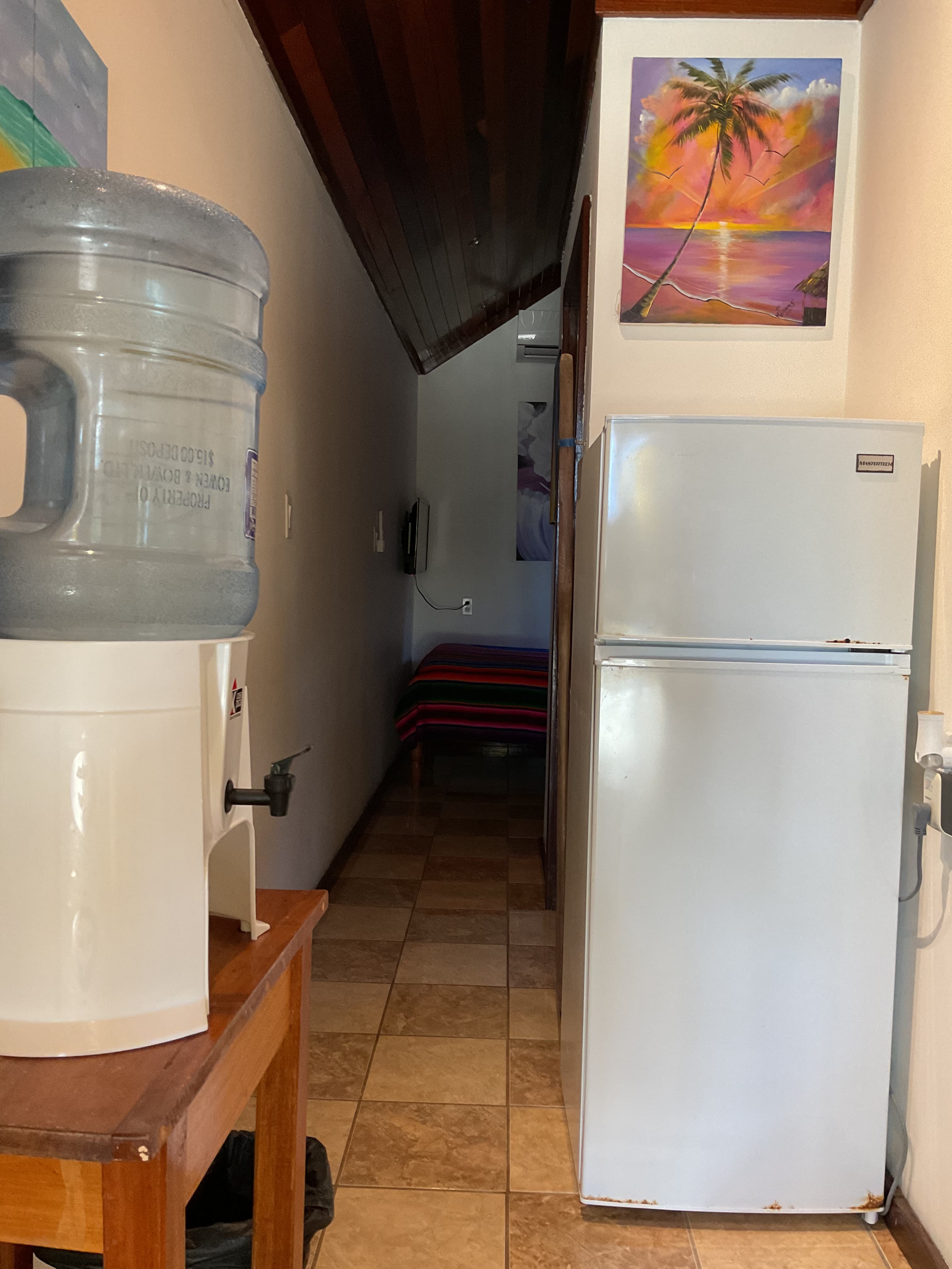 A hallway featuring a water cooler, a white refrigerator, and a colorful palm tree painting.