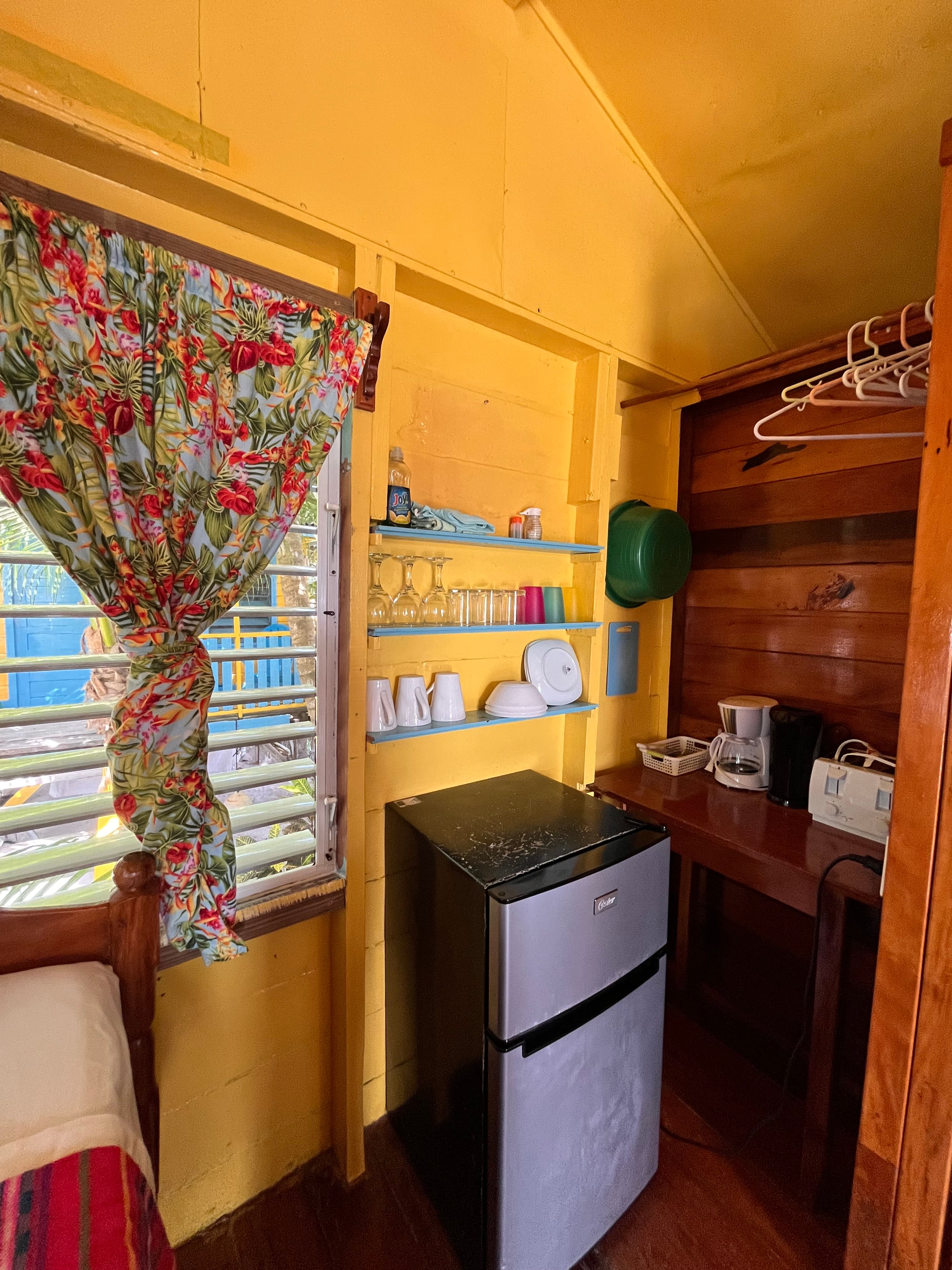 A cozy interior of a bright yellow room featuring a mini fridge, a wooden table with a coffee maker, and a floral curtain by a window.