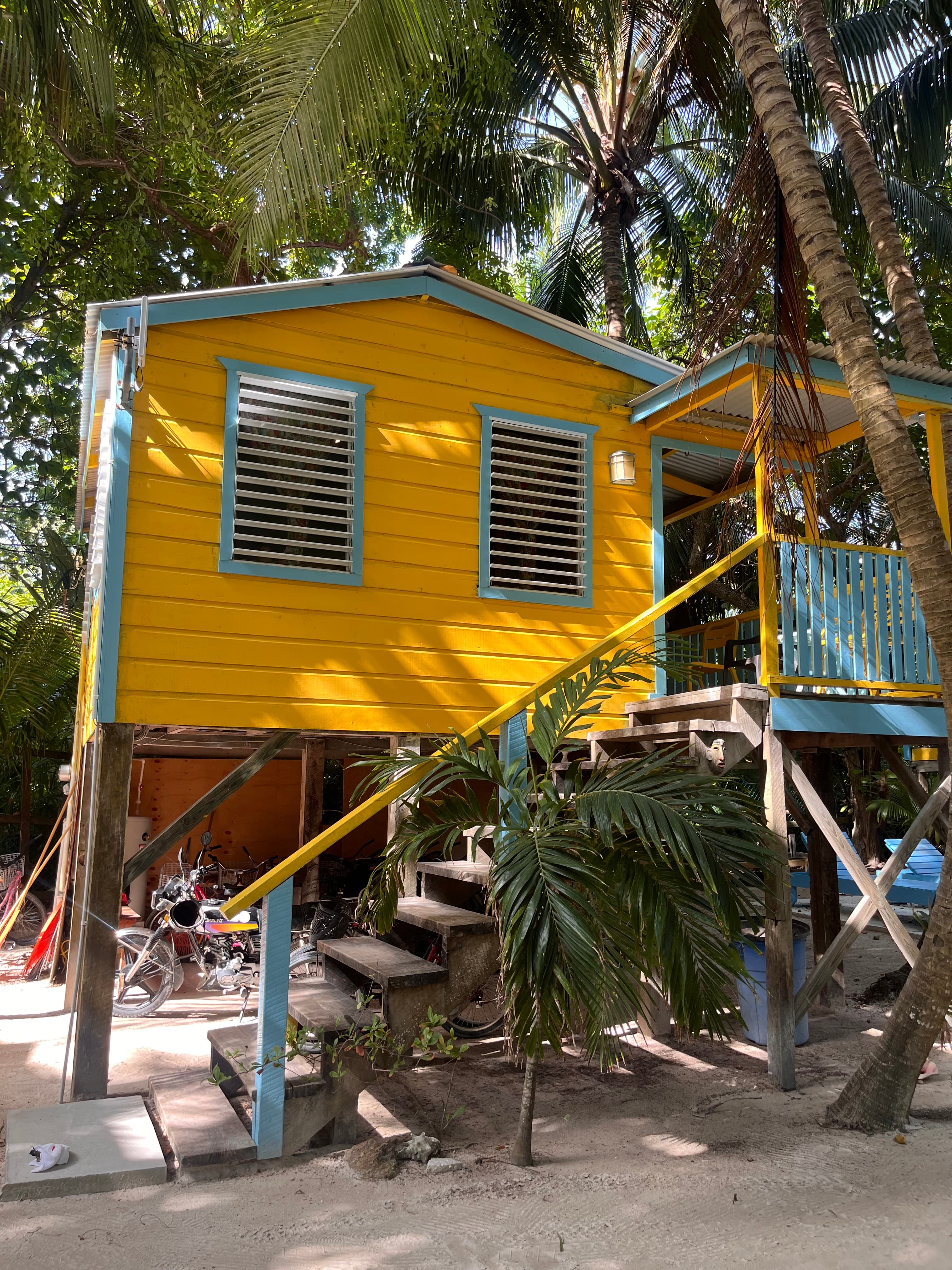 A bright yellow wooden house elevated on stilts, surrounded by palm trees.