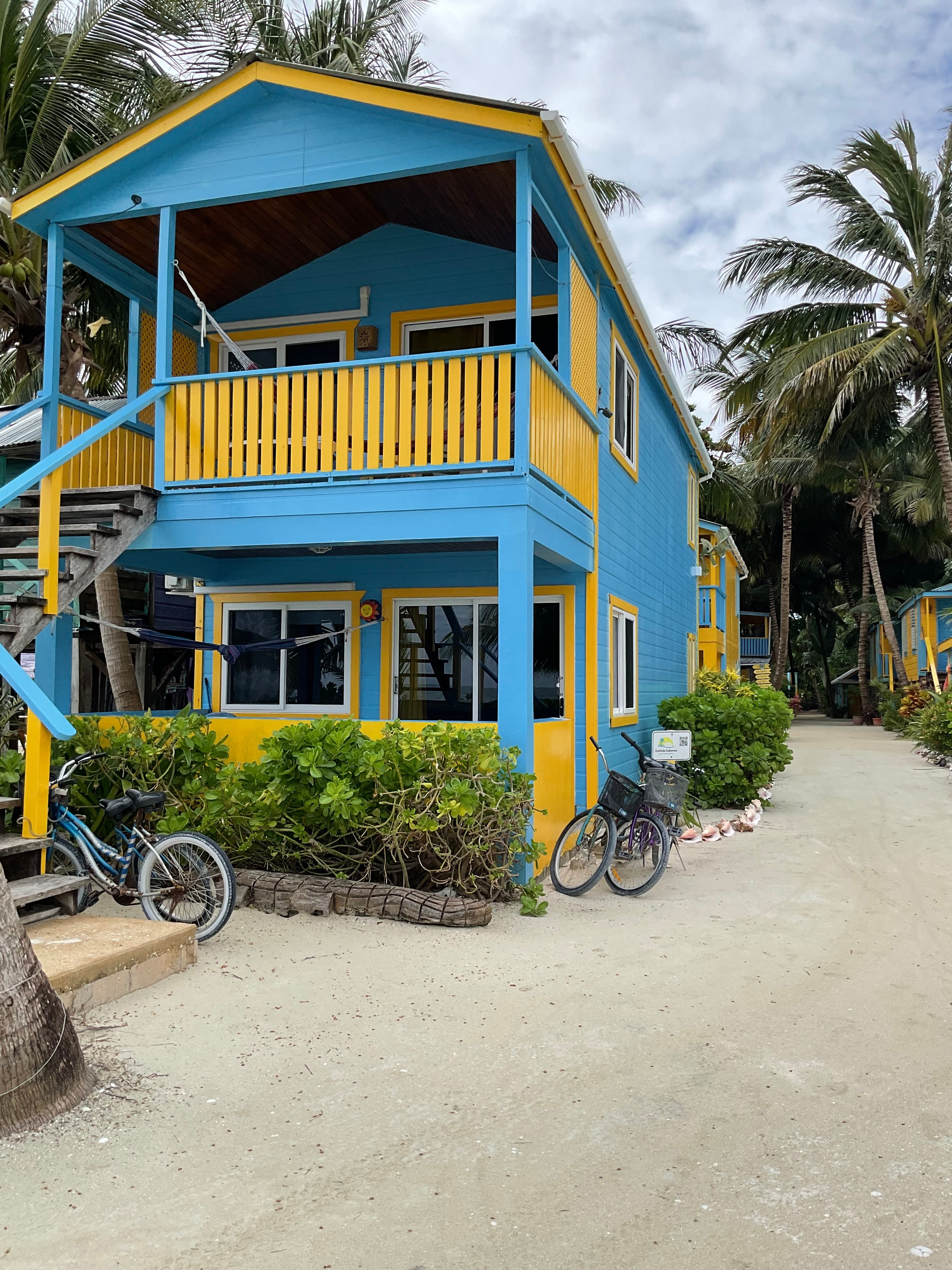 A colorful blue and yellow two-story house surrounded by palm trees and bicycles on a sandy pathway.