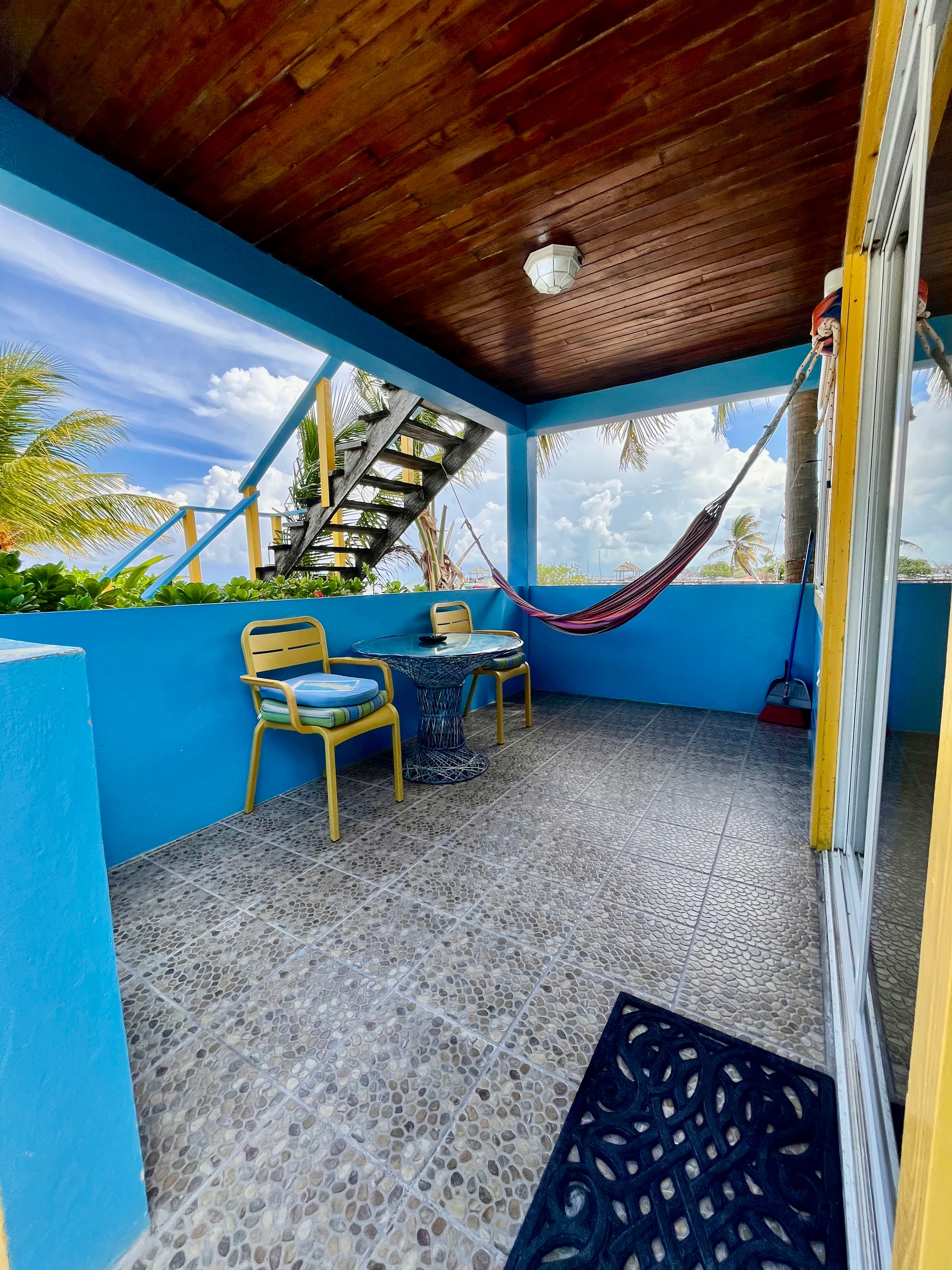 A cozy outdoor seating area featuring blue walls, two chairs, a small table, and a hammock, with stairs leading up and a view of palm trees and clouds.